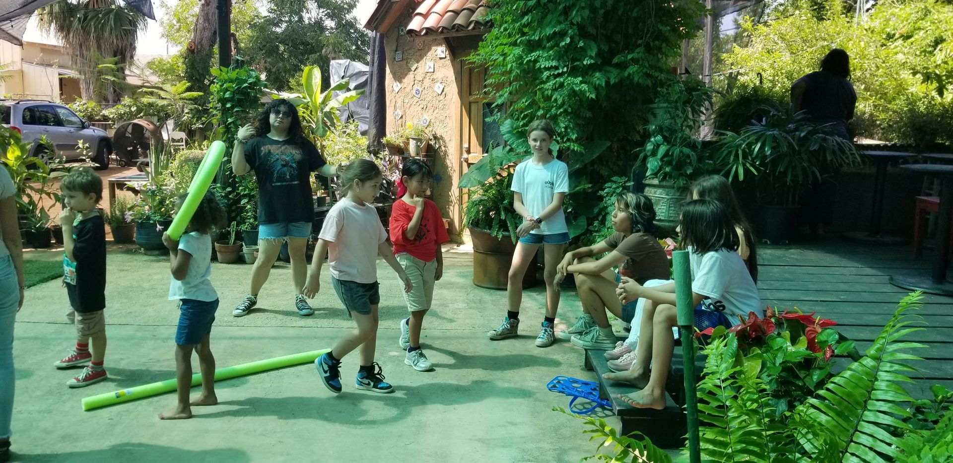 Children playing outside with pool noodles. Some are sitting, others are walking or standing.