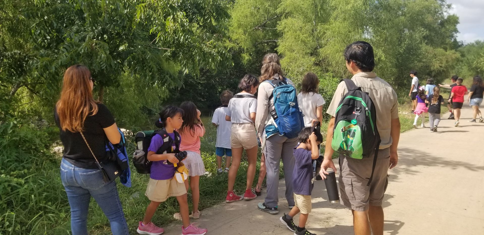 Group of children and adults walking along a path next to green trees on a sunny day.