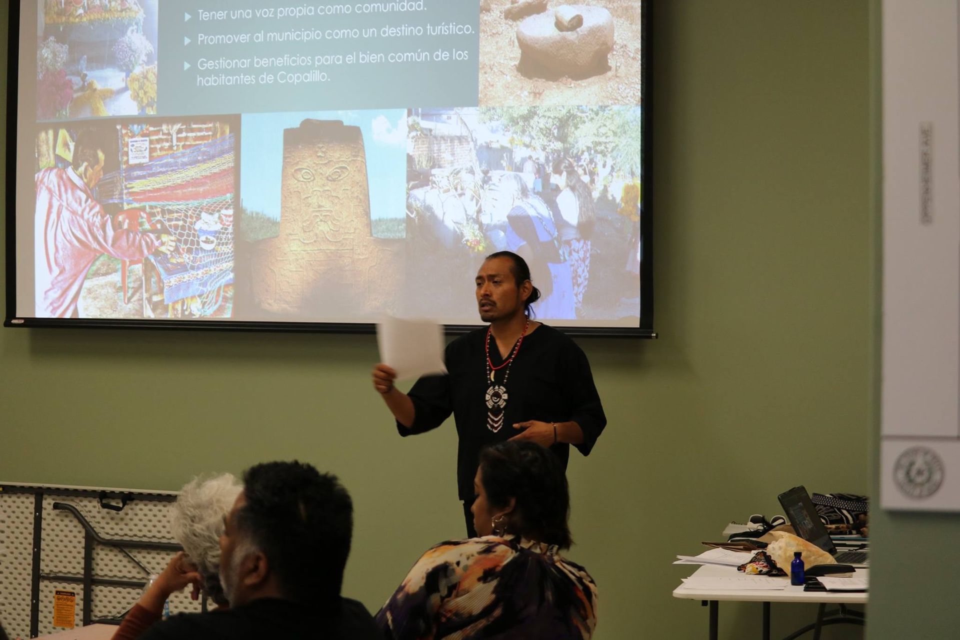 Man giving presentation; projected images, audience in room.