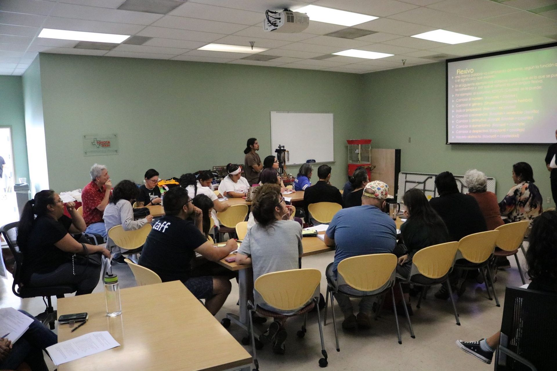 A group of people in a meeting room, watching a presentation on a screen.