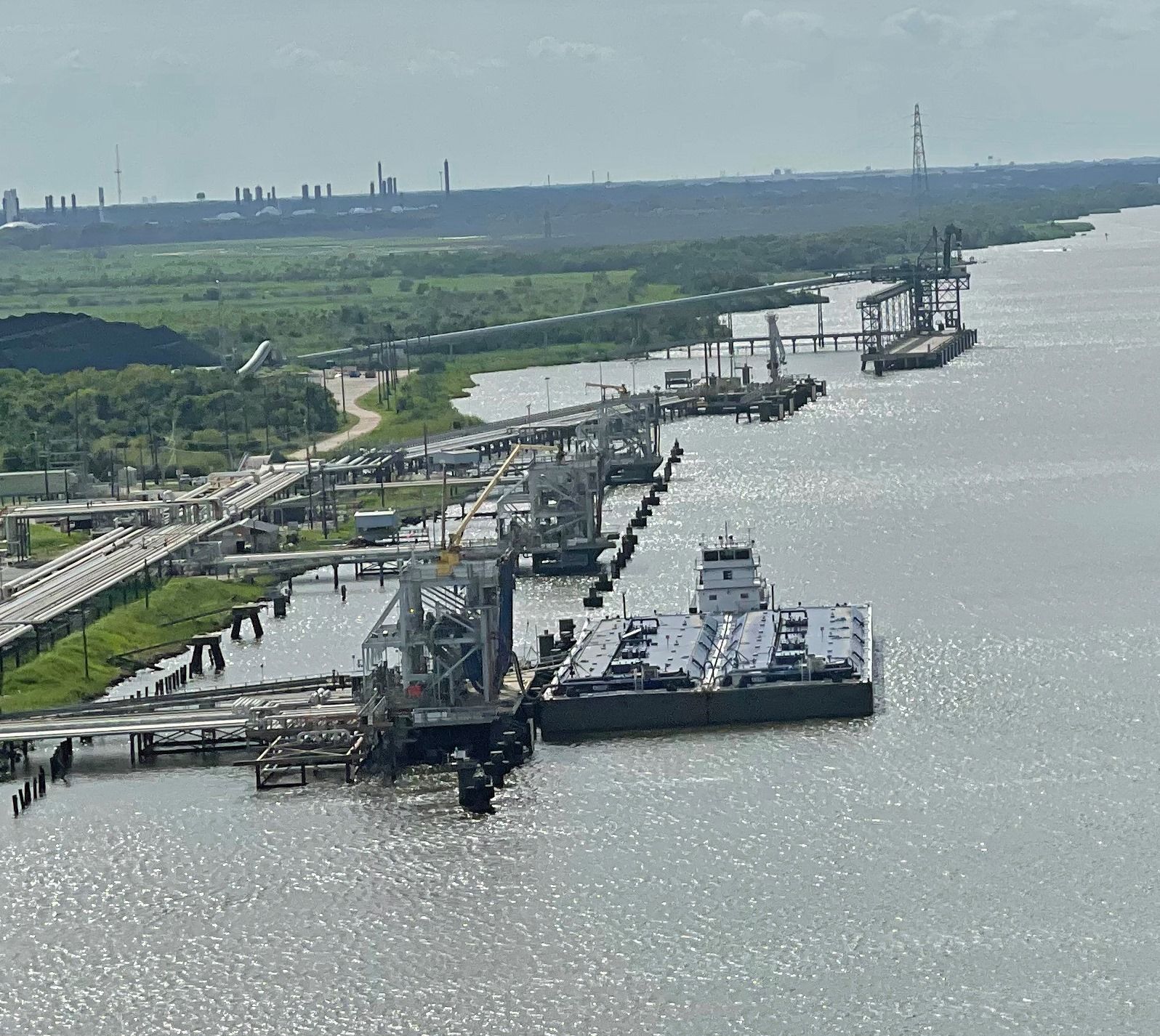 A barge docks near industrial structures on a river, with a bridge and distant factories.