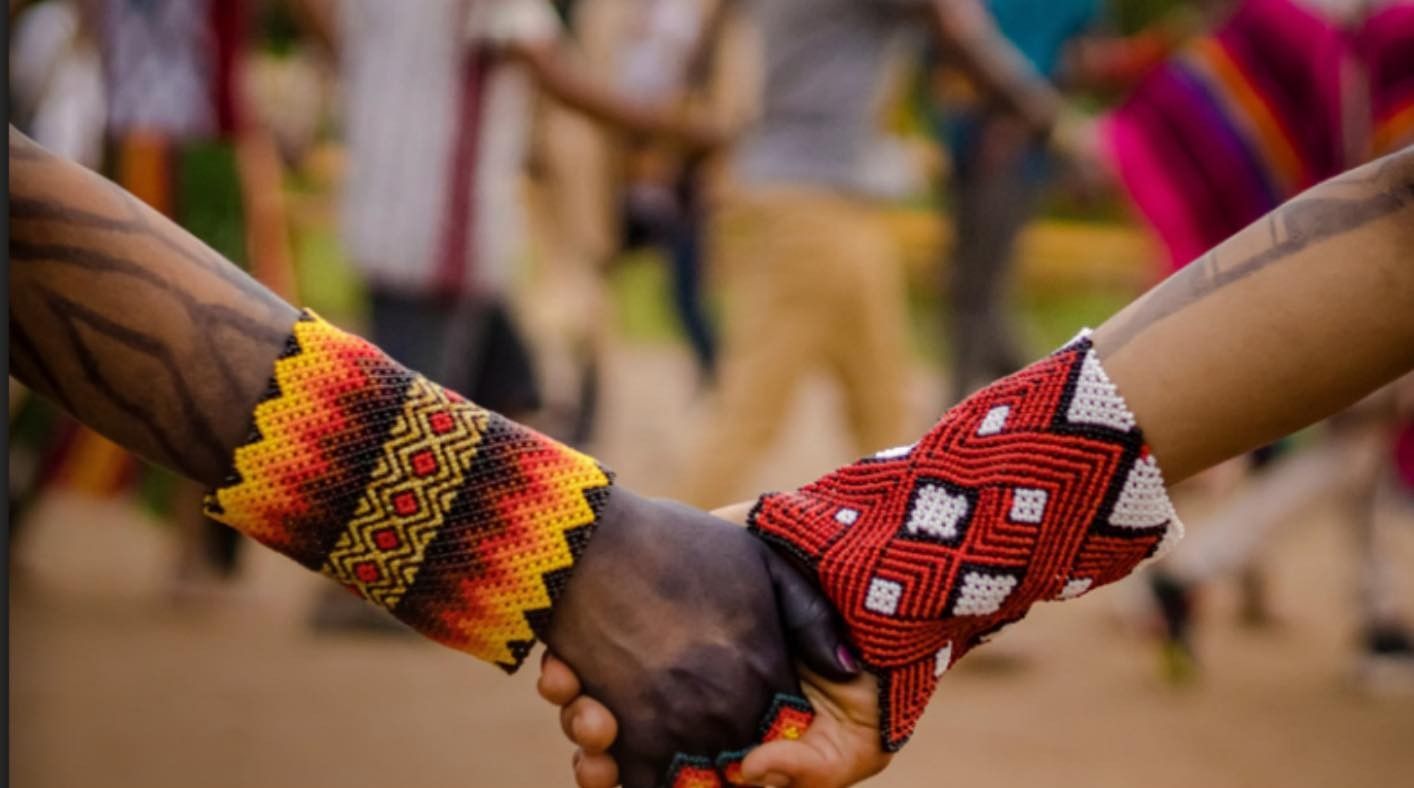 Two hands clasped together, adorned with beaded bracelets. People in background.