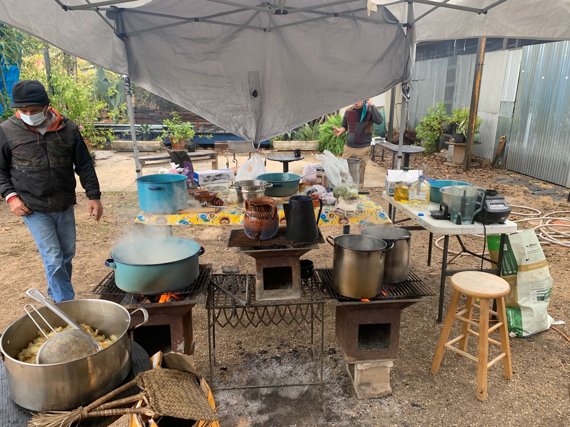 People cooking outdoors over open fires under a tent. Pots and food on tables. One man wears a mask.