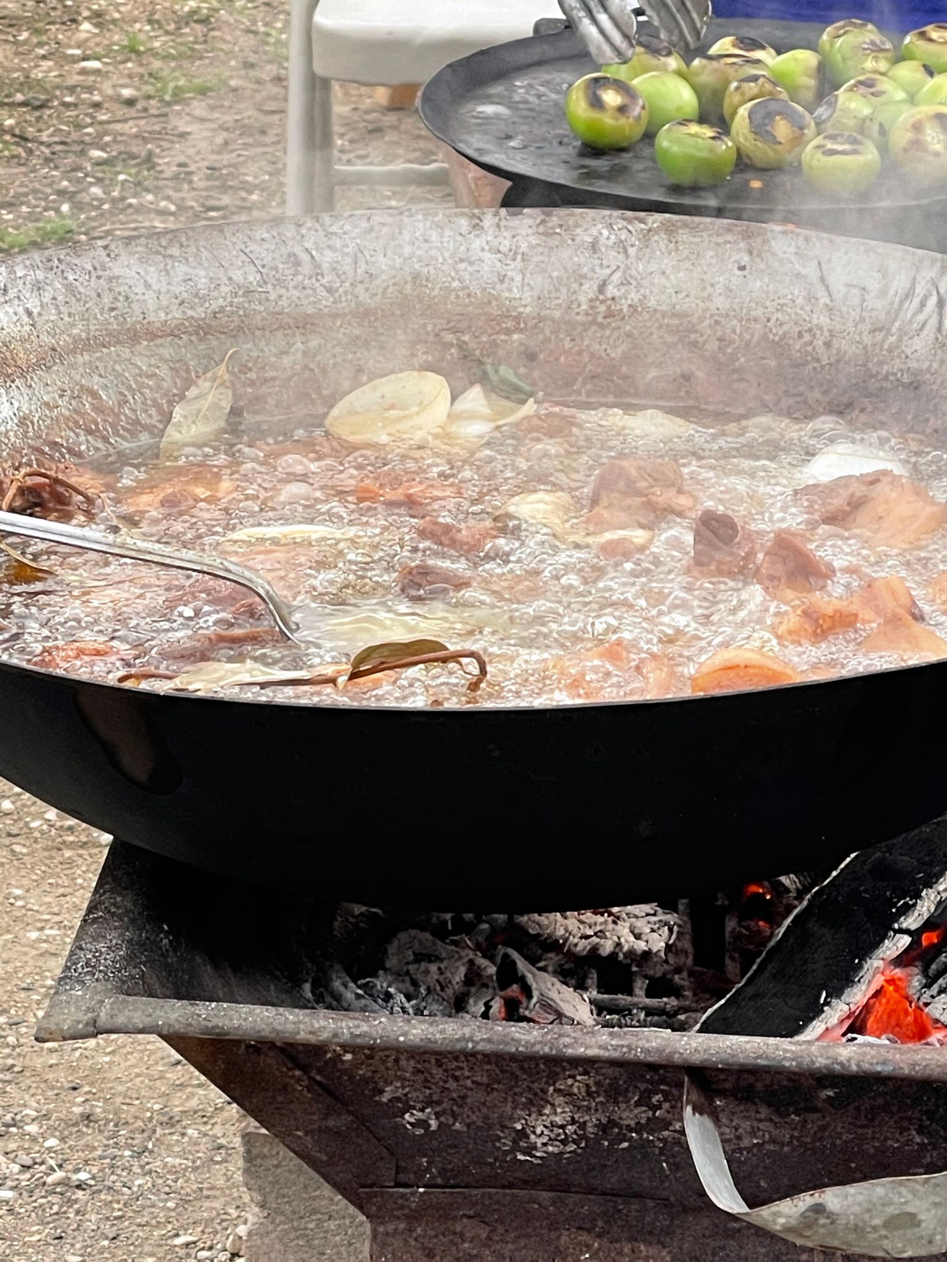 Large pot of food cooking over open flames. Green tomatillos are in a pan.