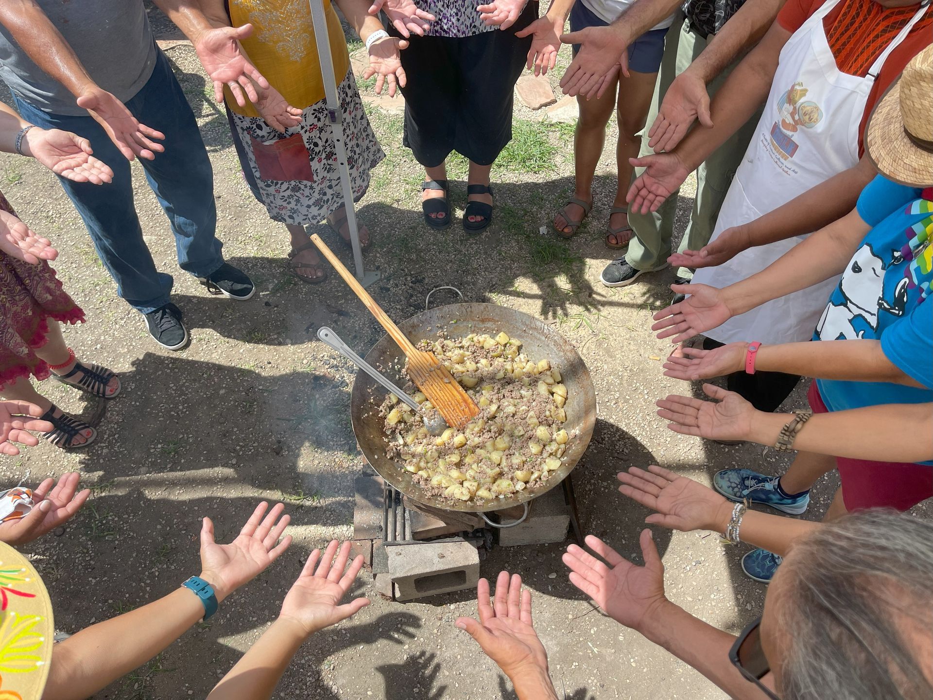 People surrounding a large outdoor cooking pot, hands outstretched.