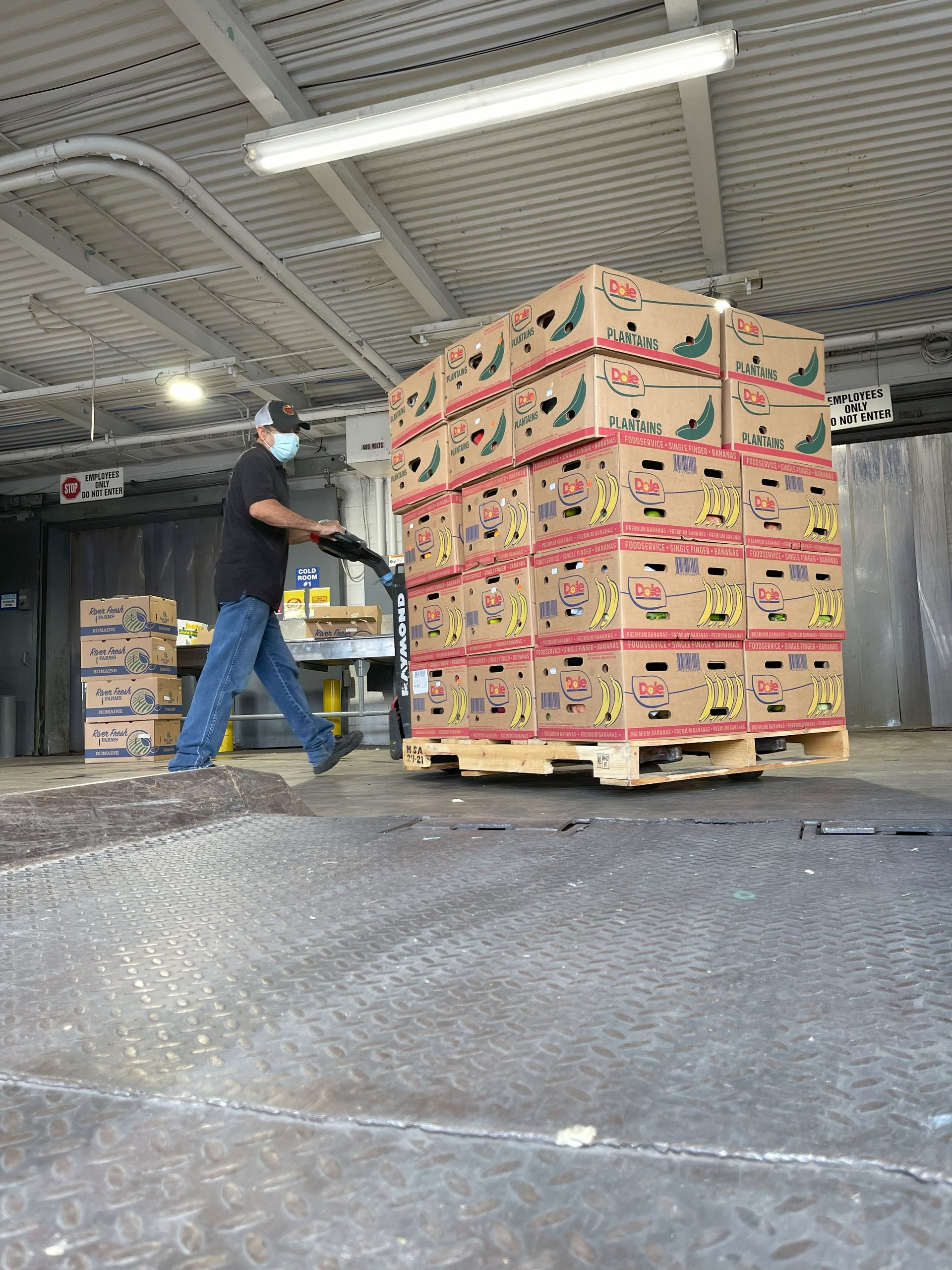 Man in mask moves stacked boxes of produce on a pallet with a pallet jack at a loading dock.