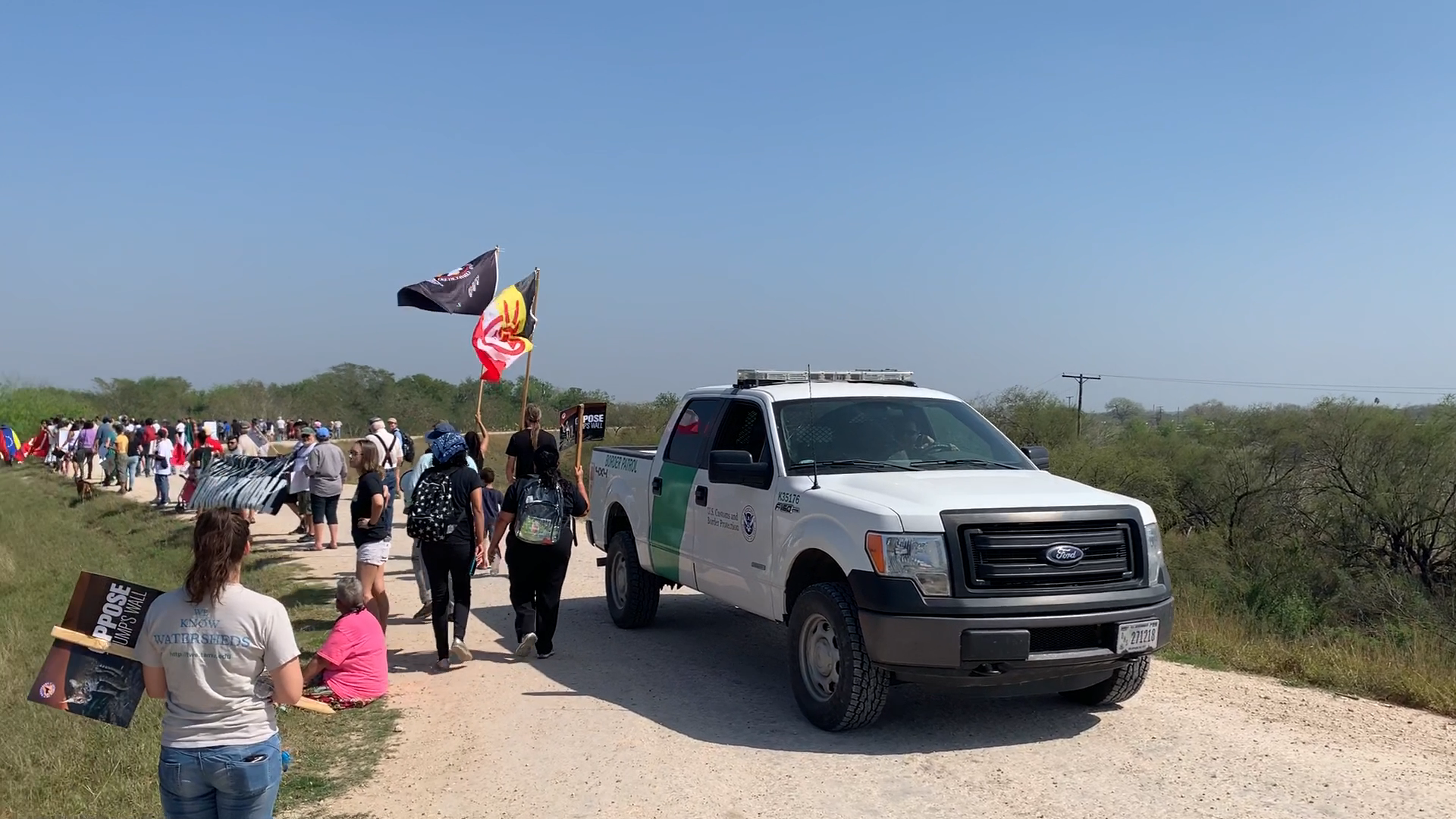 People walking on a dirt road along the border, with Border Patrol truck.