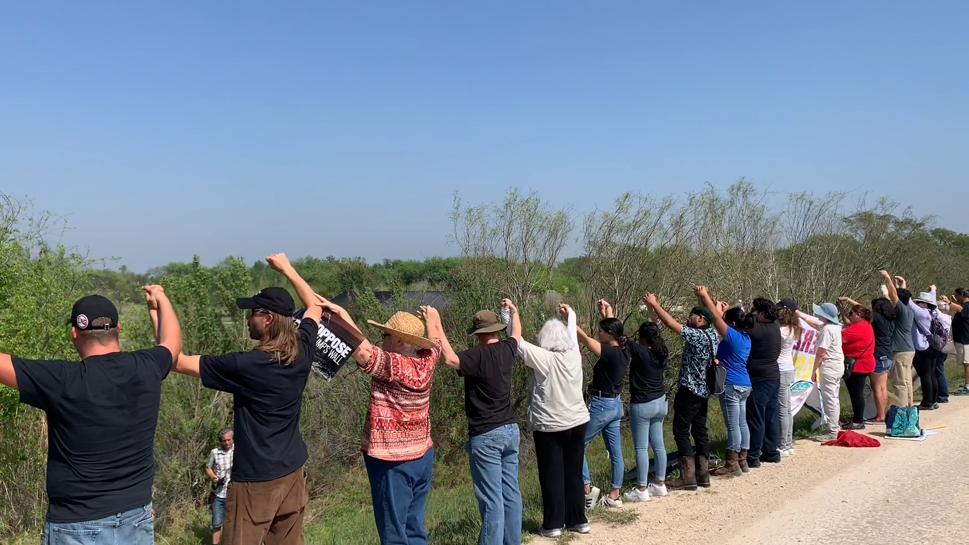A line of people with raised arms outdoors, a mix of ages and clothing, facing a green landscape.