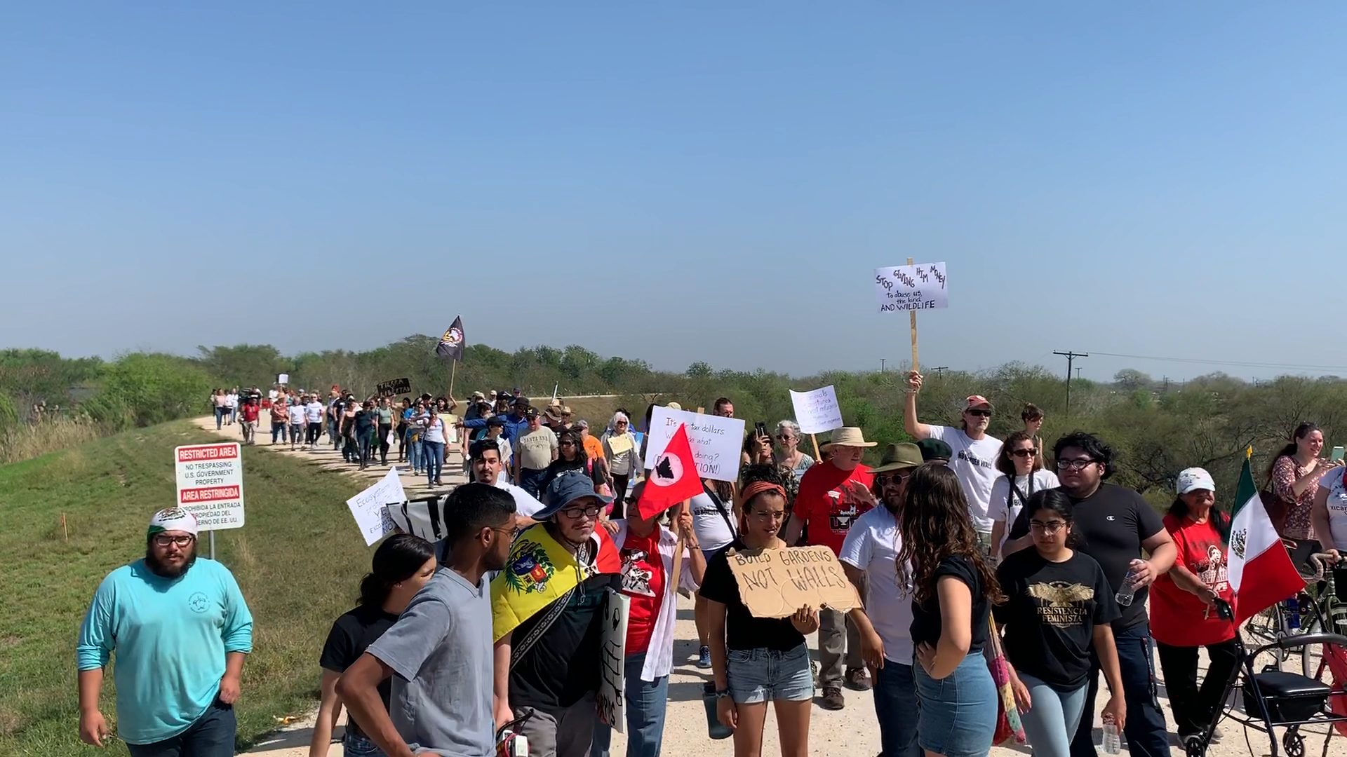 People marching on a path, holding signs and flags, possibly a protest in daylight.