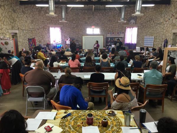 A large meeting room with people seated at tables, listening to a speaker at the front.