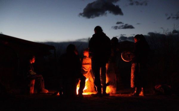 People gathered around a campfire at dusk. Silhouettes of people, orange firelight, and dark blue sky.