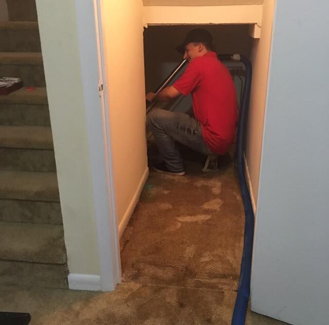 Person in a red shirt crouches in a hallway doorway, using a tool on a pipe or cable.