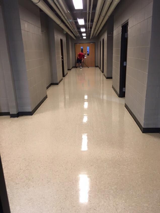 Long beige hallway with gray walls and bright ceiling lights; two people stand near the far end.