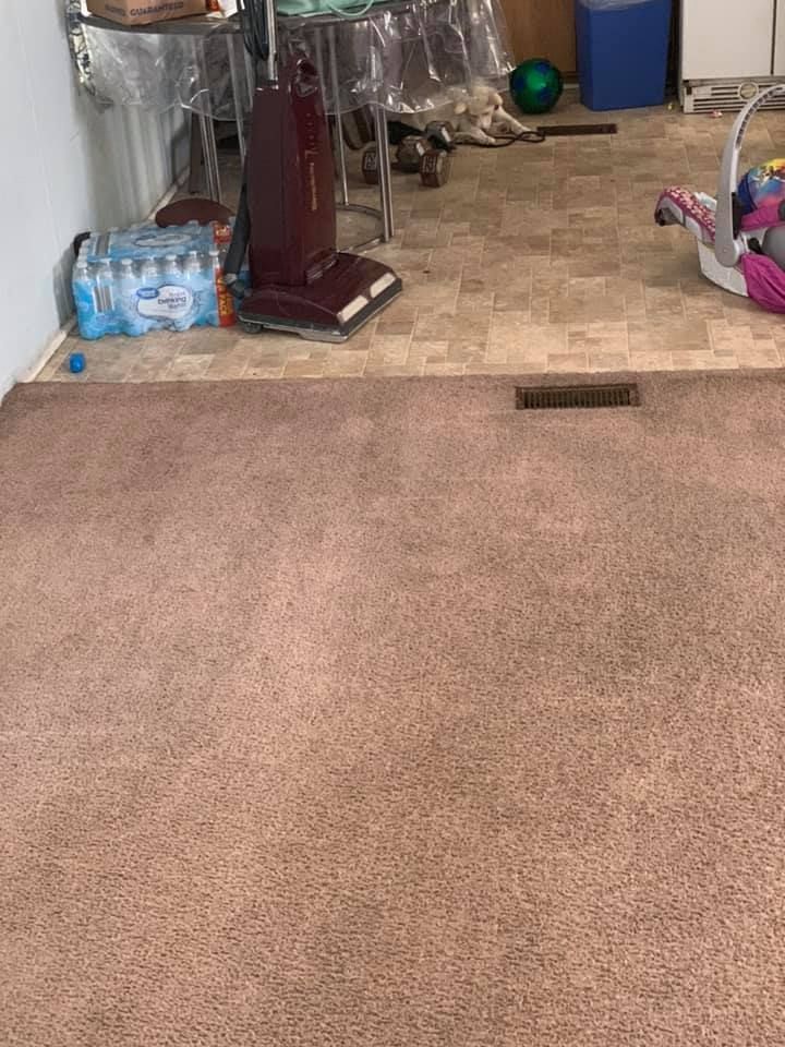 Carpeted floor with a rectangular floor vent, vacuum, and water bottles near a tiled area