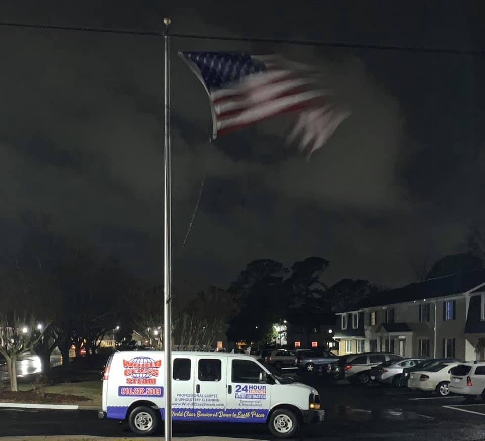 White service van parked at night beneath a large waving American flag on a tall pole.