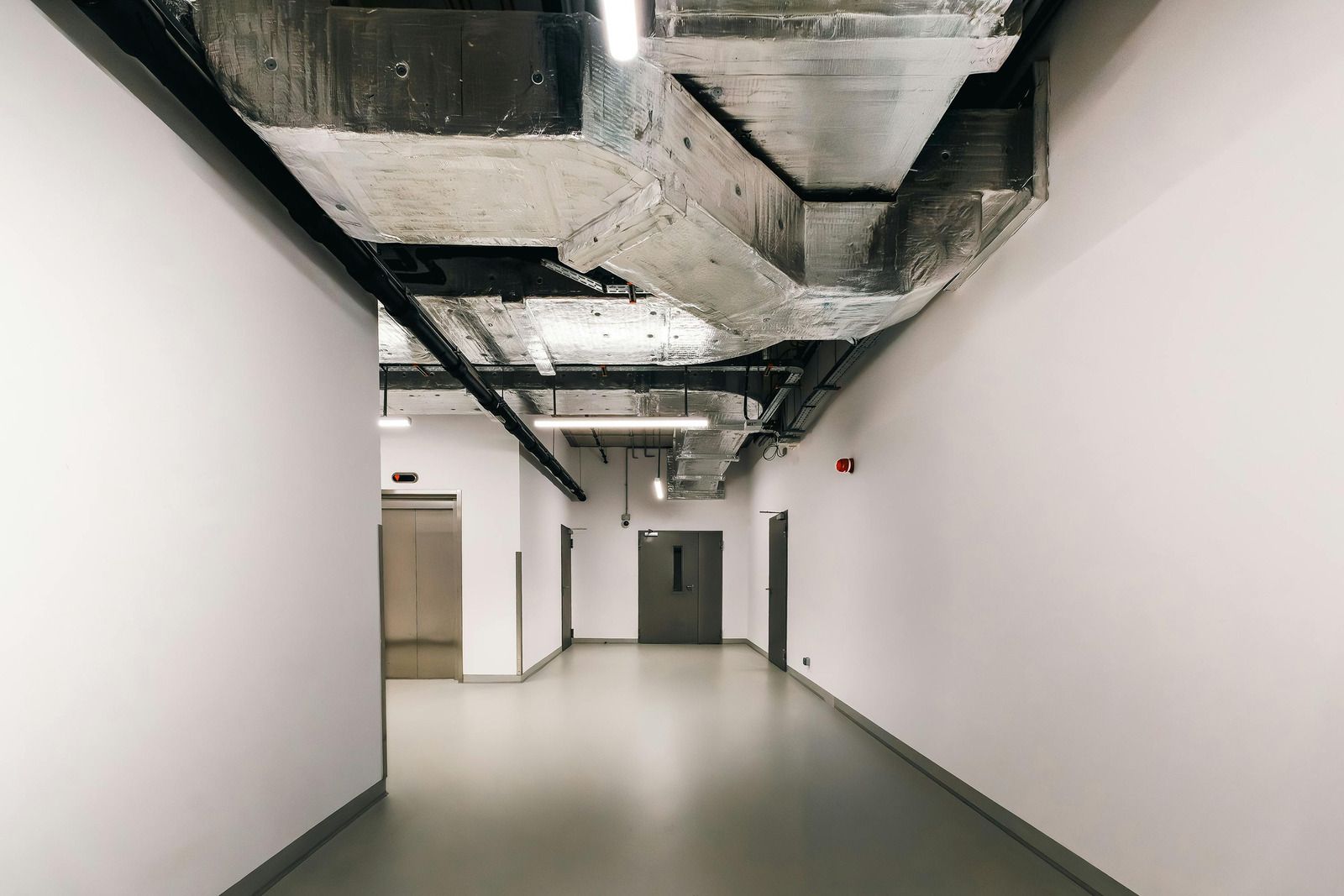 Long hallway with white walls and exposed concrete ceiling, leading to closed doors.