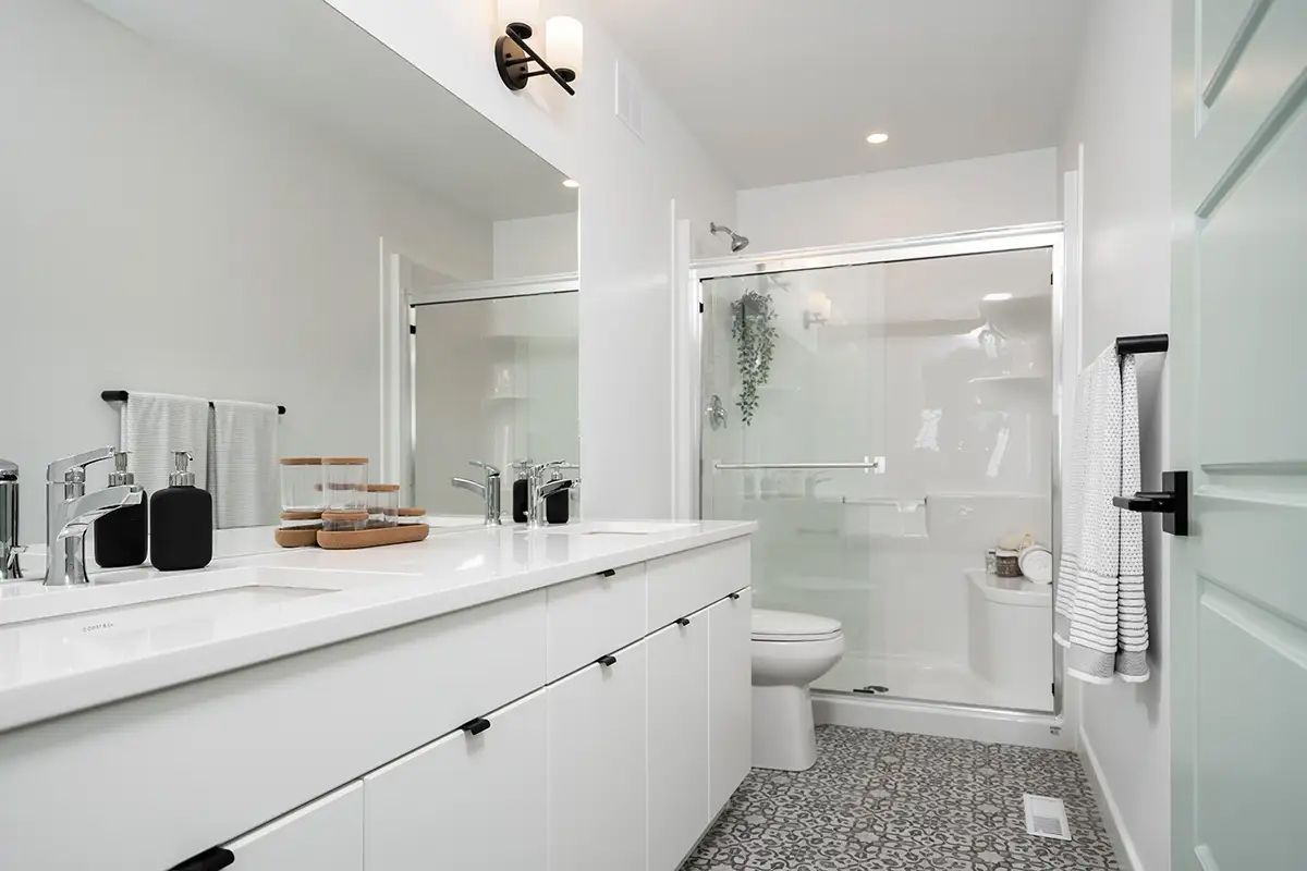 White bathroom with dual sinks, shower, and patterned floor.