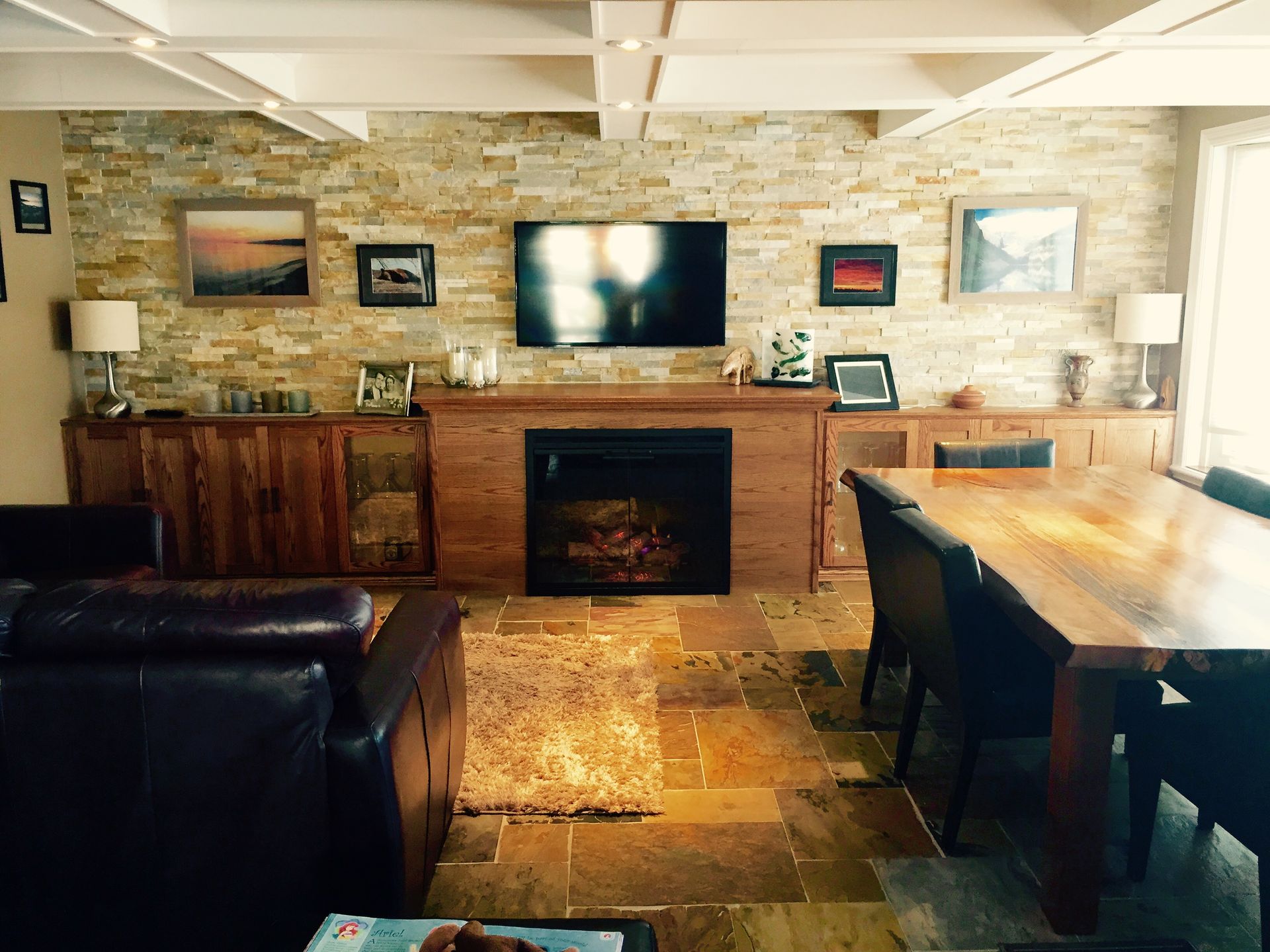 Living room with stone wall, fireplace, TV, wood cabinets, dining table, leather sofa, and artwork.