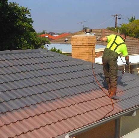 A Man is Painting the Roof of a House with a Sprayer — Roof Repairs in Maitland, NSW
