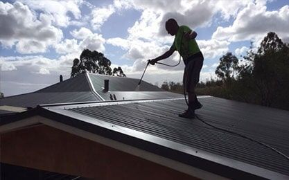 A Man is Cleaning the Roof of a House with a High Pressure Washer — Roof Repairs in Maitland, NSW