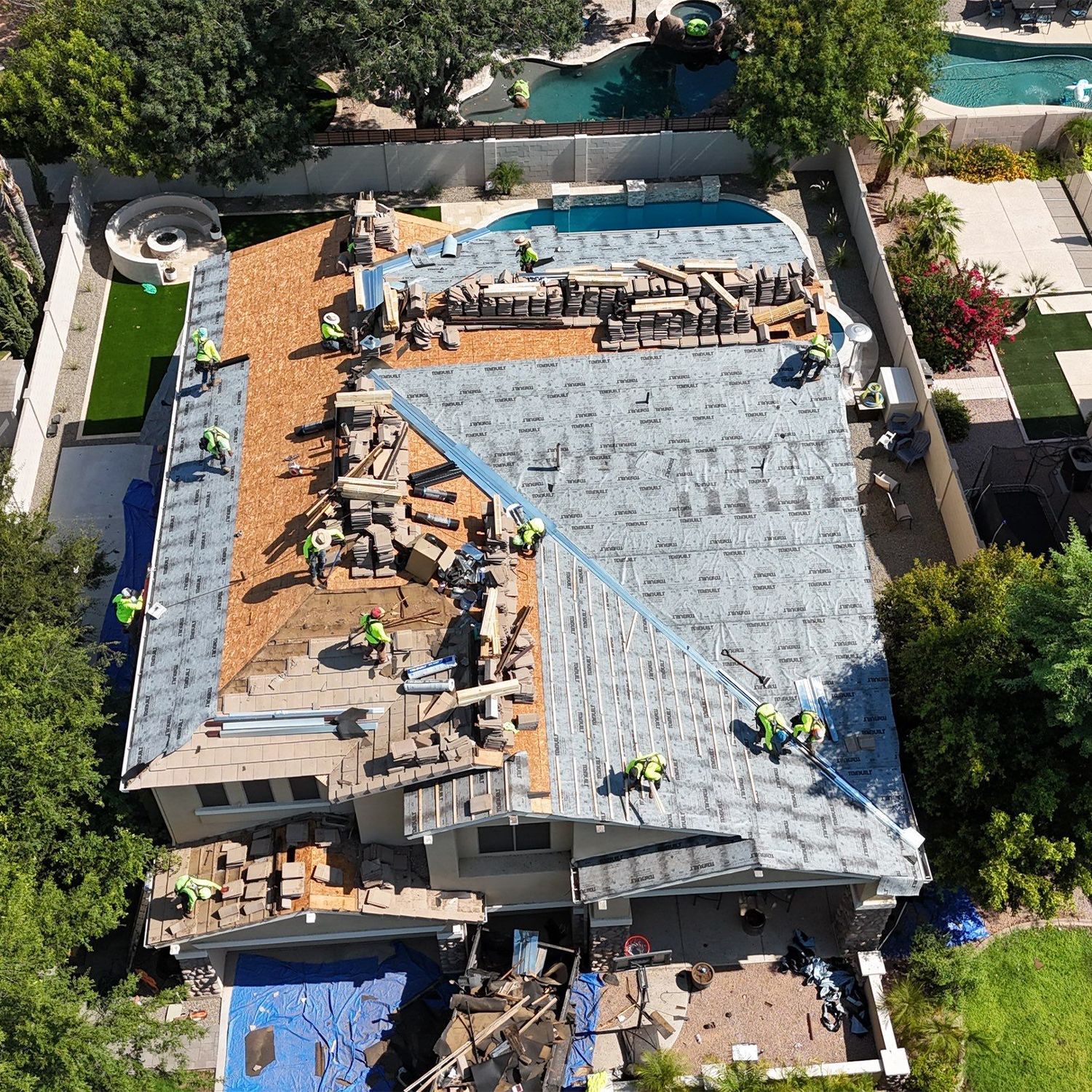 Aerial view of a house with roof construction in progress. Workers install roofing materials; blue tarp covers some areas.