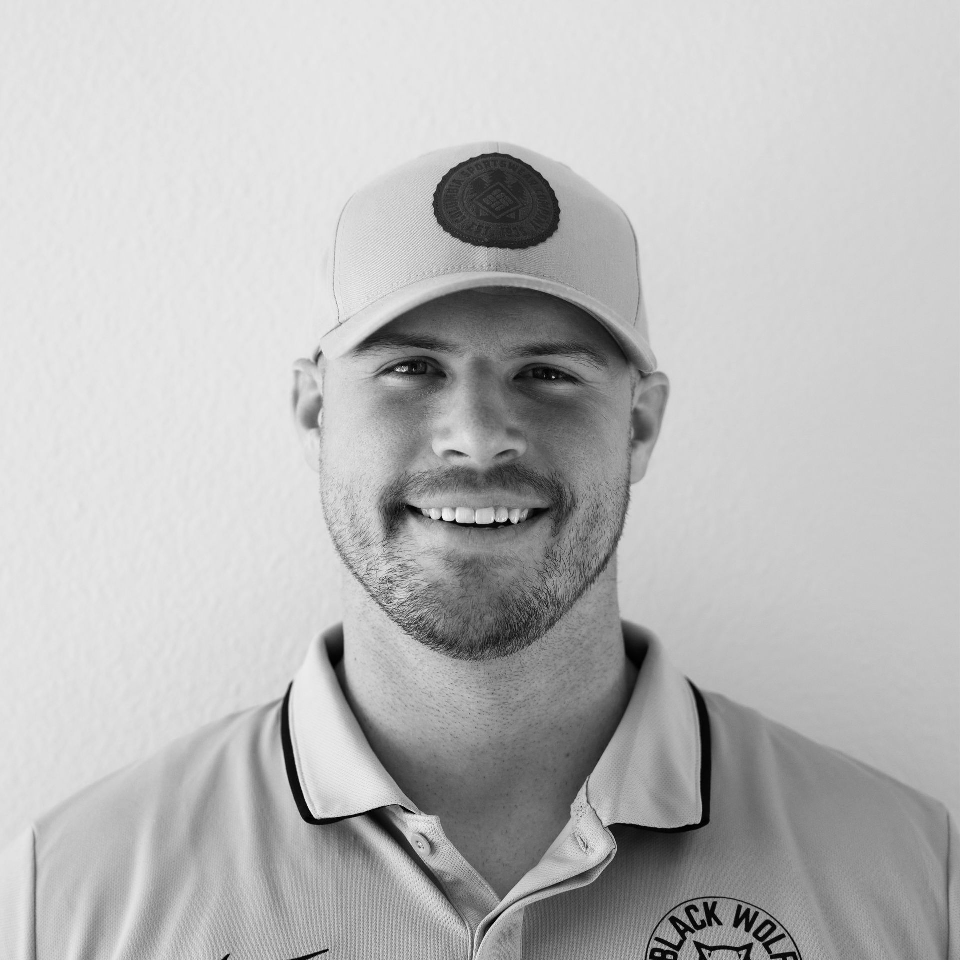 Man smiling, wearing a baseball cap with a circle emblem and a collared shirt, in front of a light background.