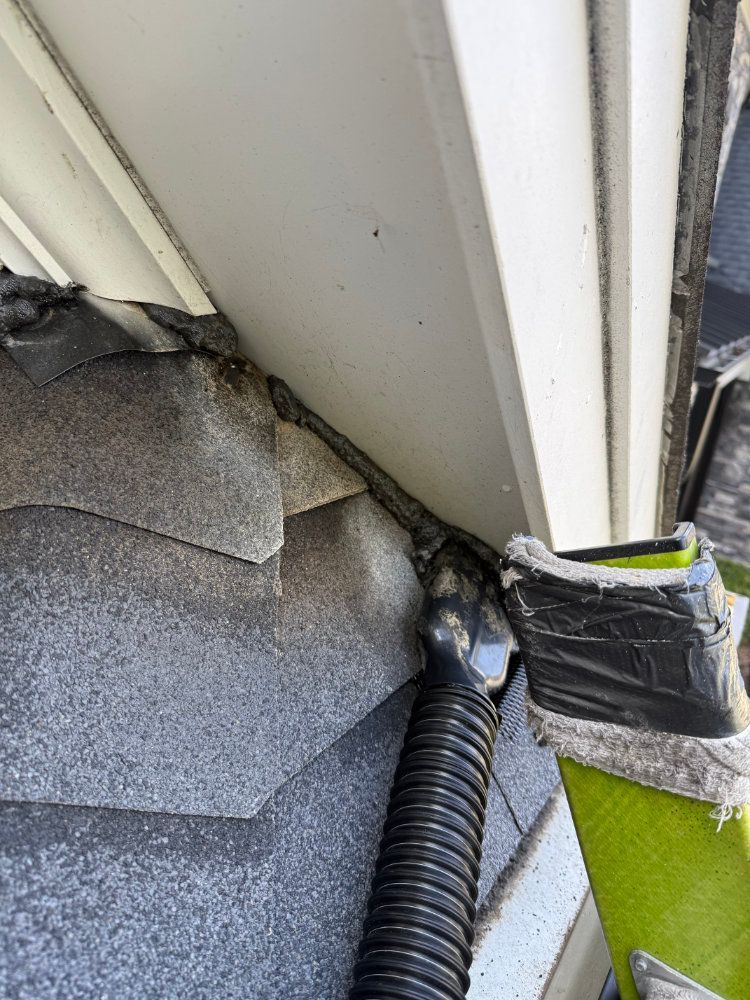 Damaged roof corner with torn shingles, exposed siding, and black drainage tubing near green trim