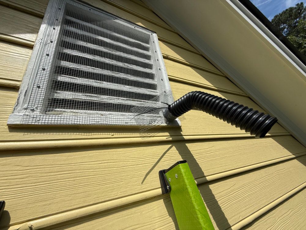 White attic vent on yellow siding with a black flexible hose and green tool attached below