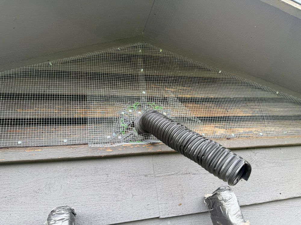 Gray corrugated hose protruding through damaged window mesh on a white wall