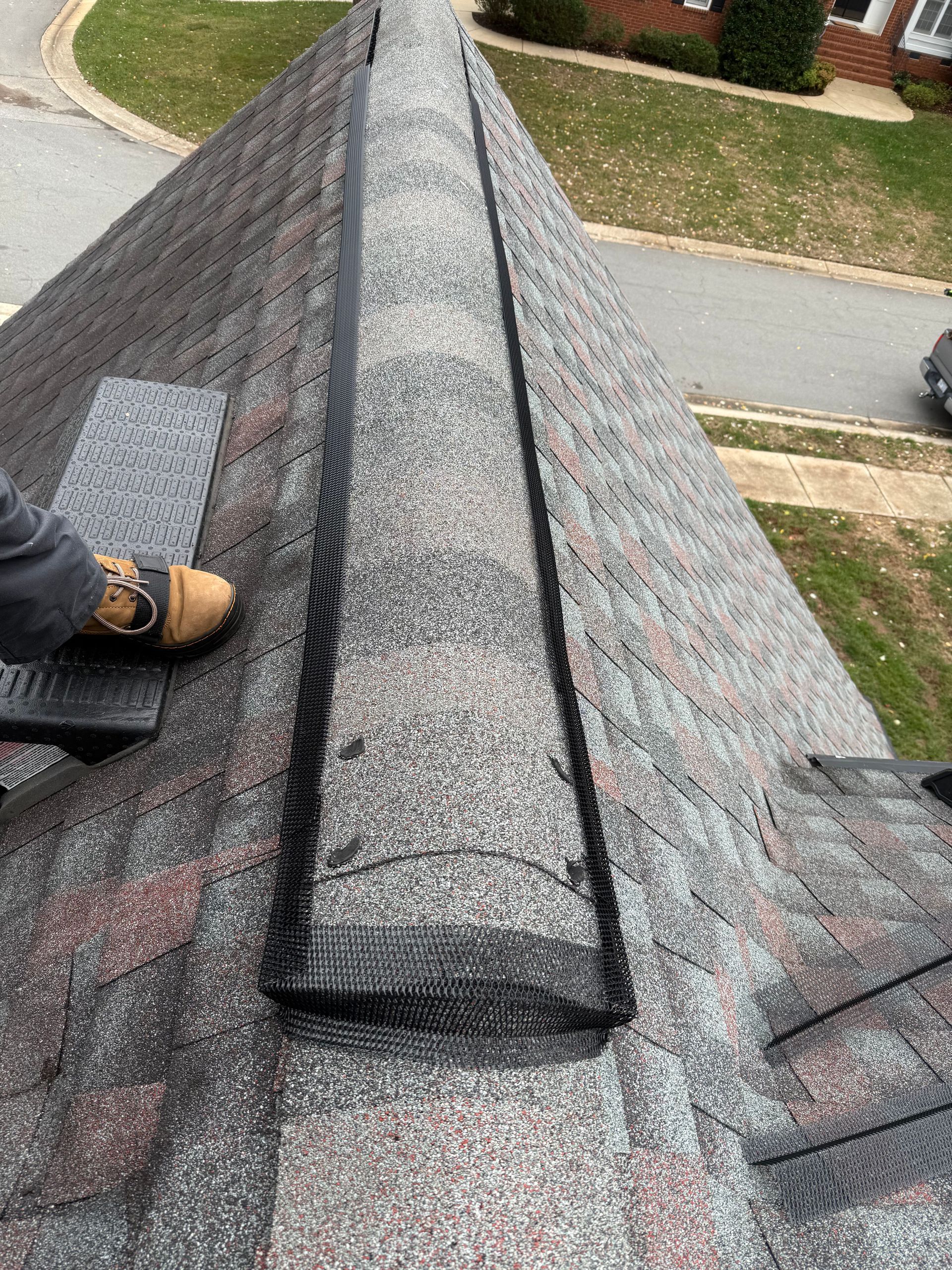 Roof ridge with gray shingles and a worker’s boot on the left side, viewed from above.
