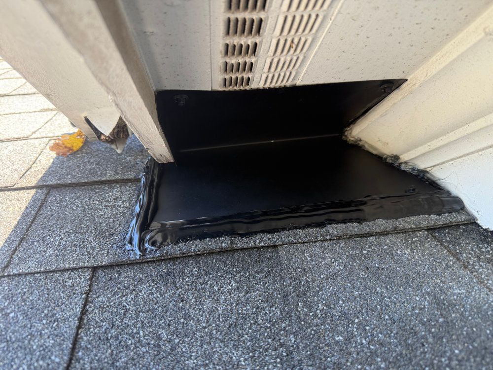 Black waterproofed vent opening on a shingled roof beside white siding, with a small leaf nearby