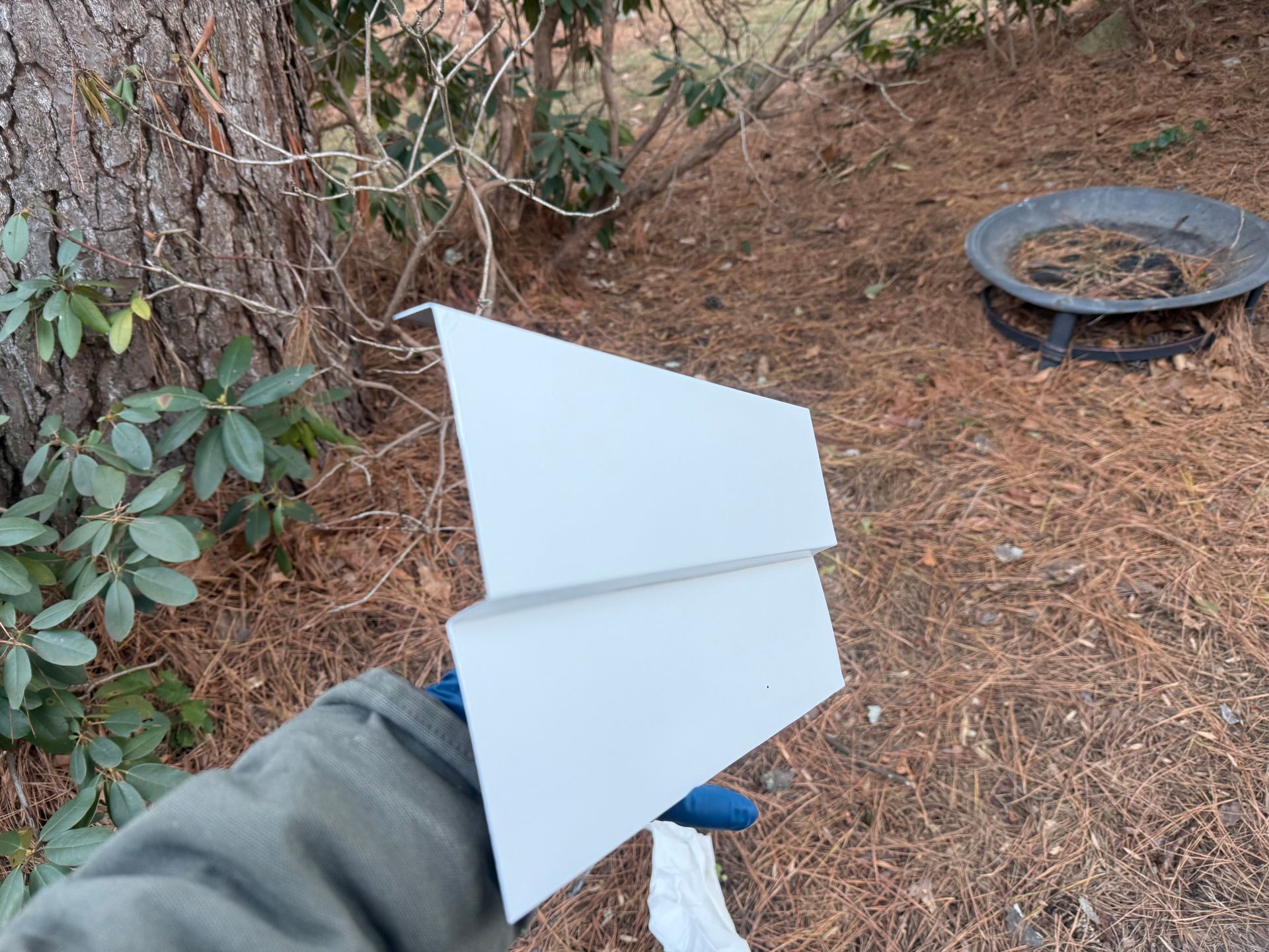 Hand holding a white paper glider outdoors near pine needles and a metal fire pit