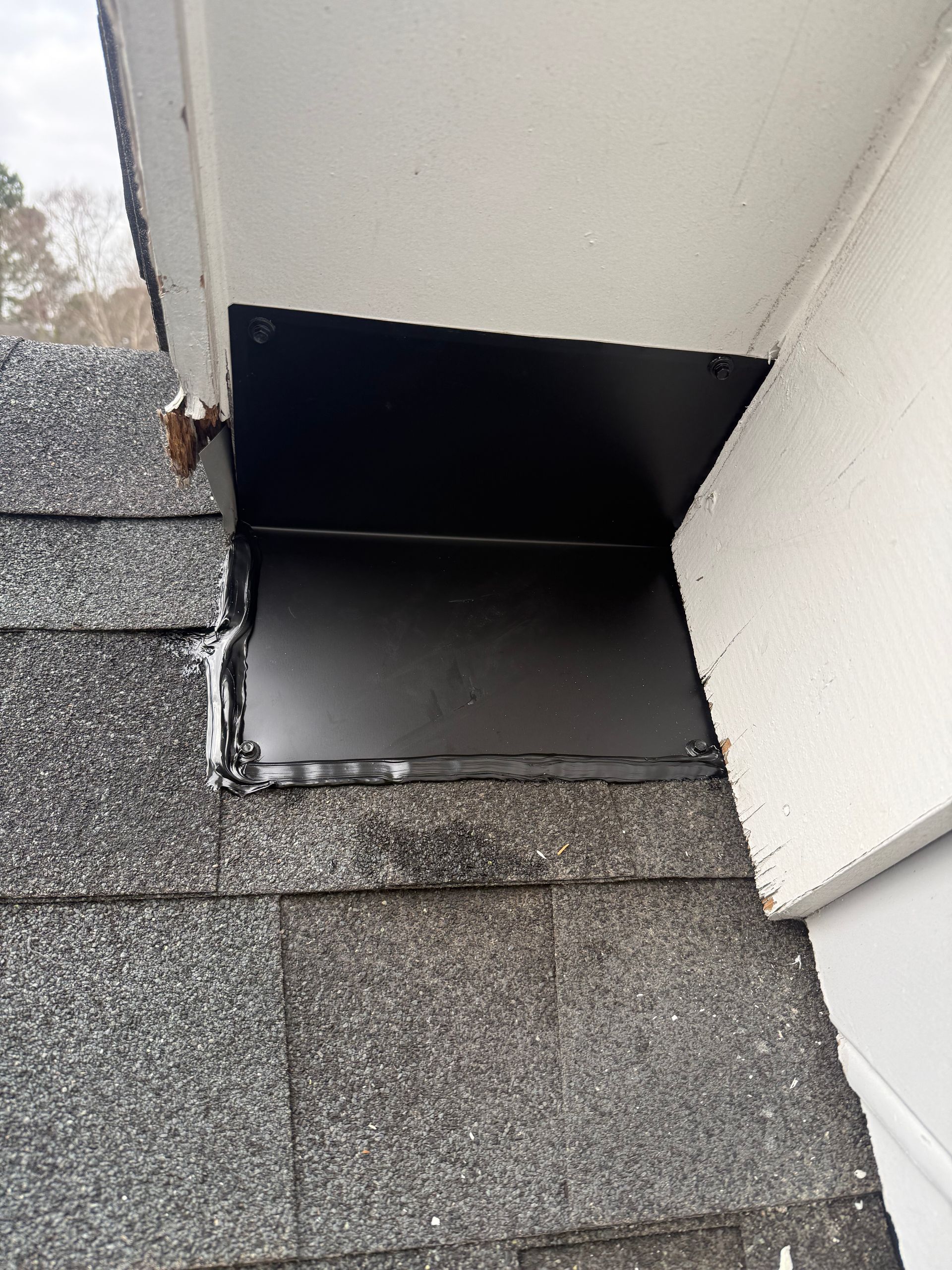 Roof vent opening with black flashing above gray shingles and white siding