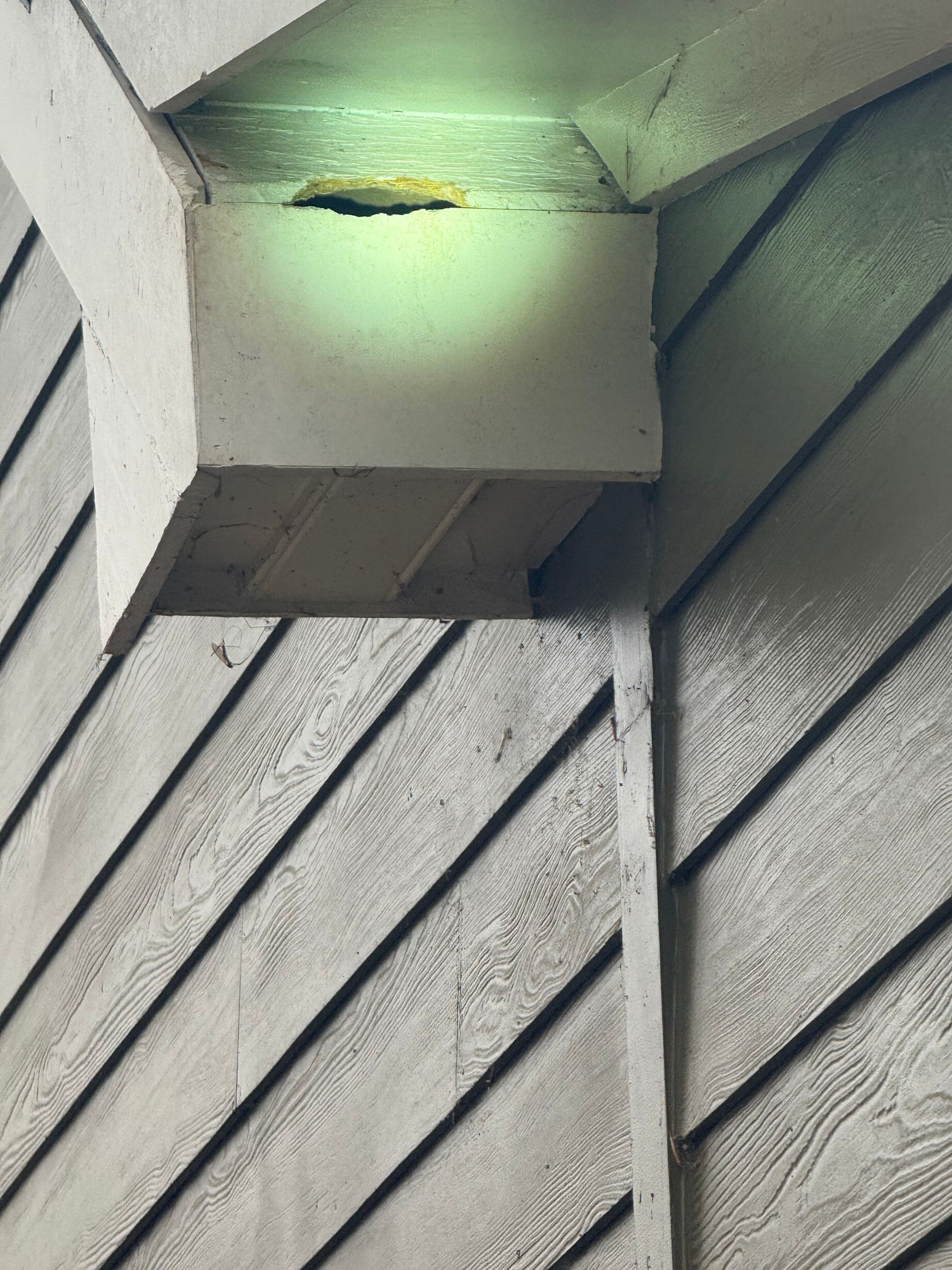 A lit exterior vent box under a roof overhang on a gray-sided building with a small hole in it