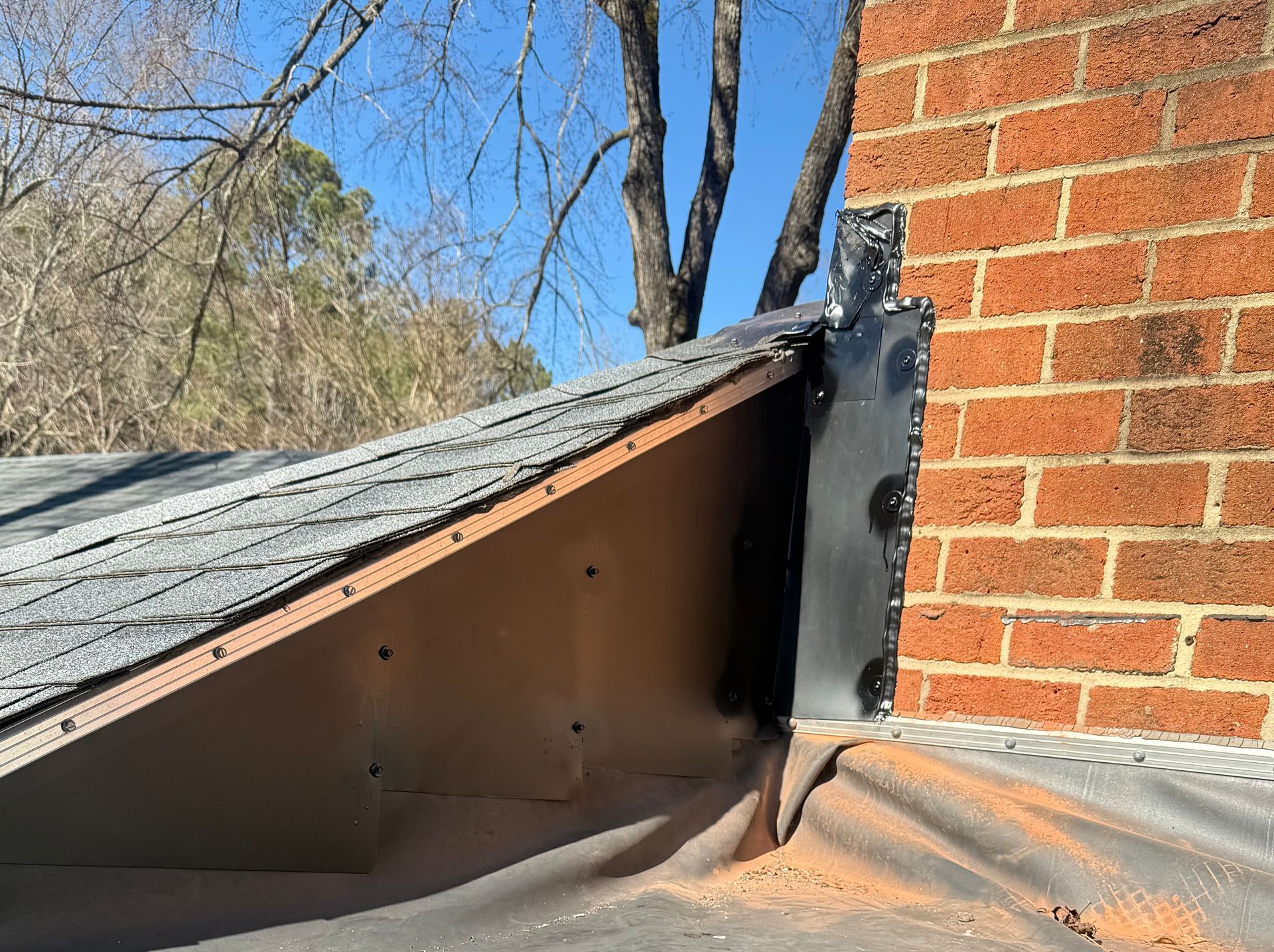 Roof flashing detail where a sloped roof meets a brick wall, with black metal trim and exposed underlayment