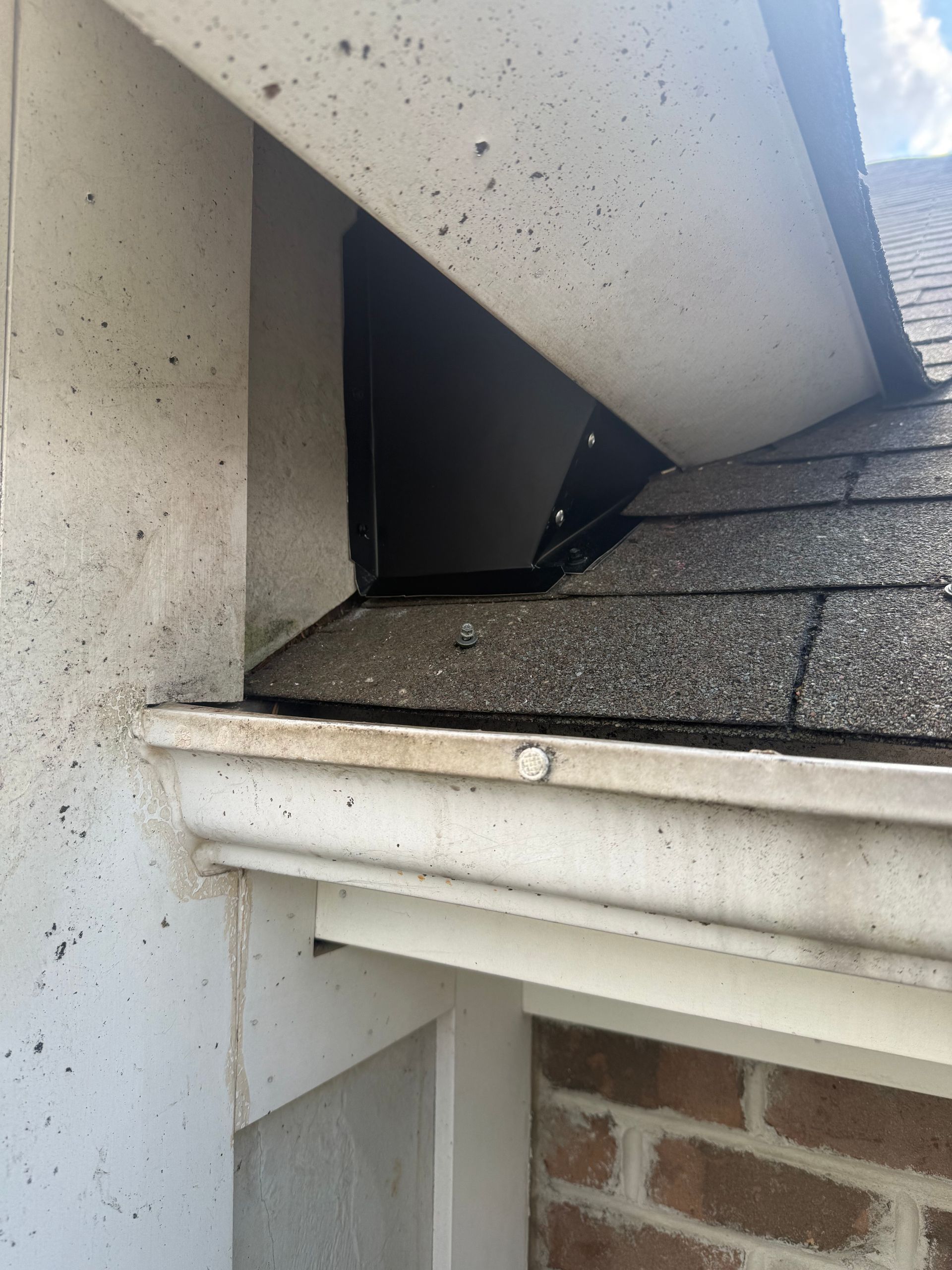 Damaged roof corner with a black opening between white trim and shingles above a brick wall.