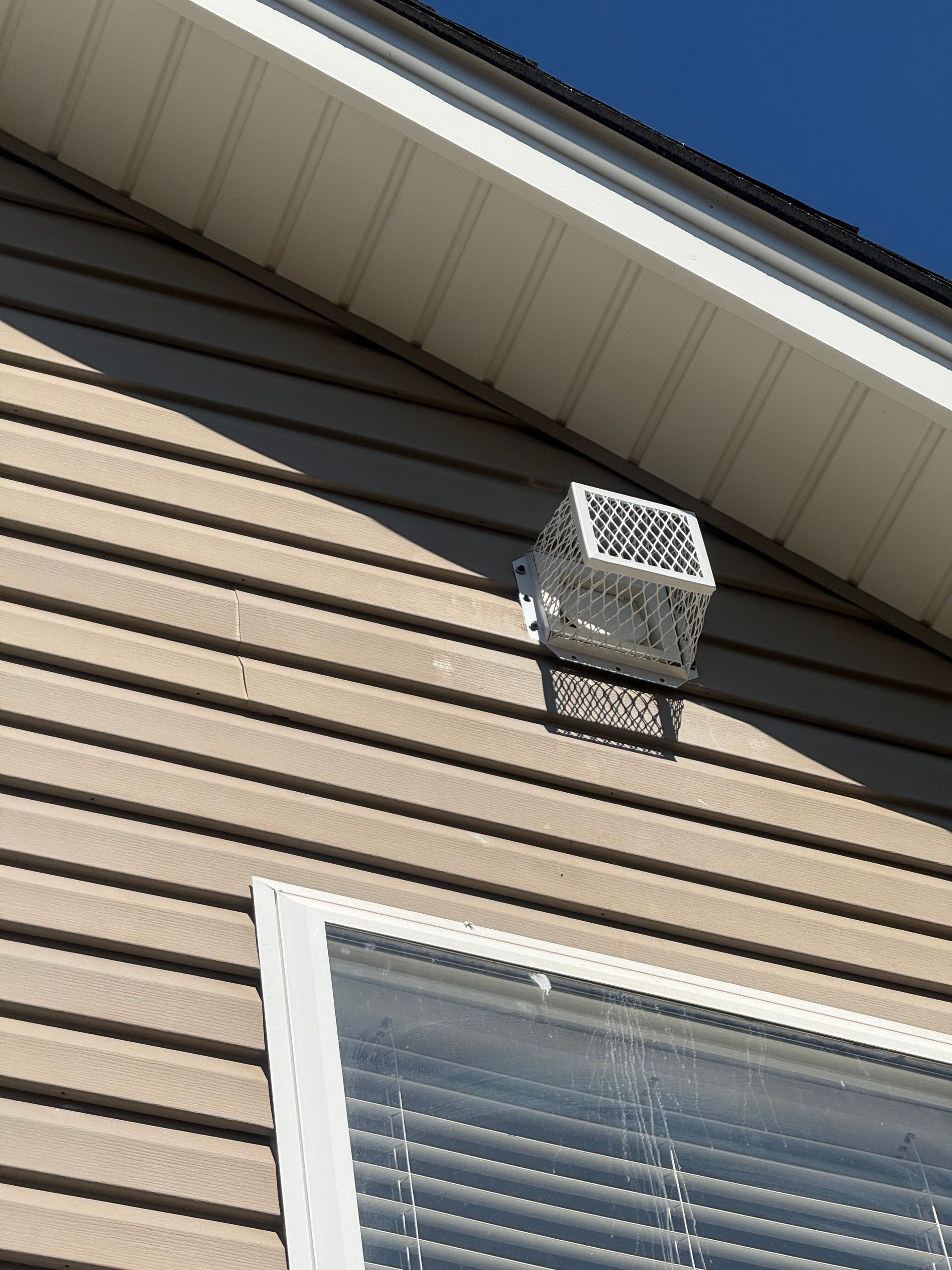 White vent on tan house siding above a window under the roof eave