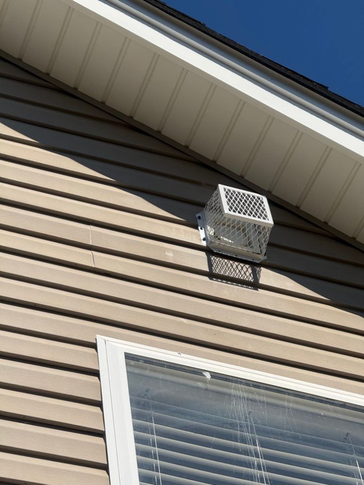 White vent box mounted on beige house siding above a window under the roof eave