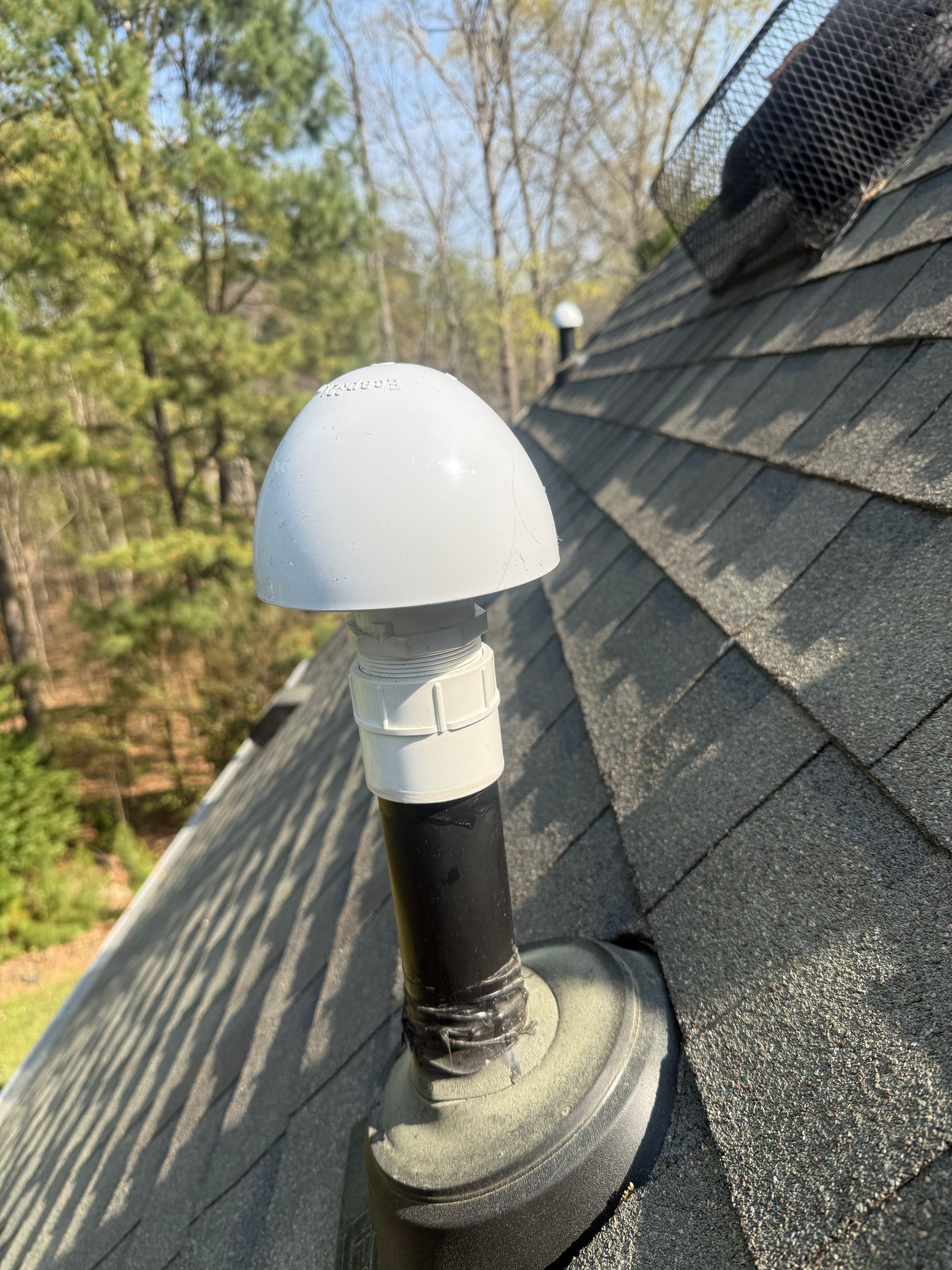 White roof vent on a shingled roof beside trees and blue sky
