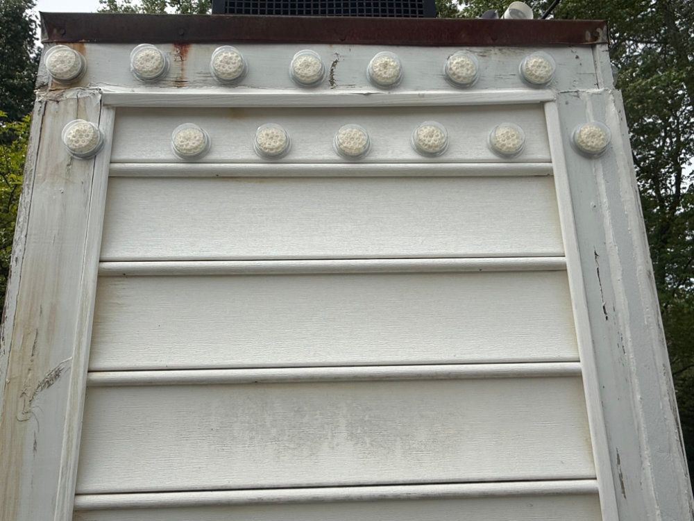 Close-up of a white decorative metal gate or vent with rivets, viewed against trees and sky.