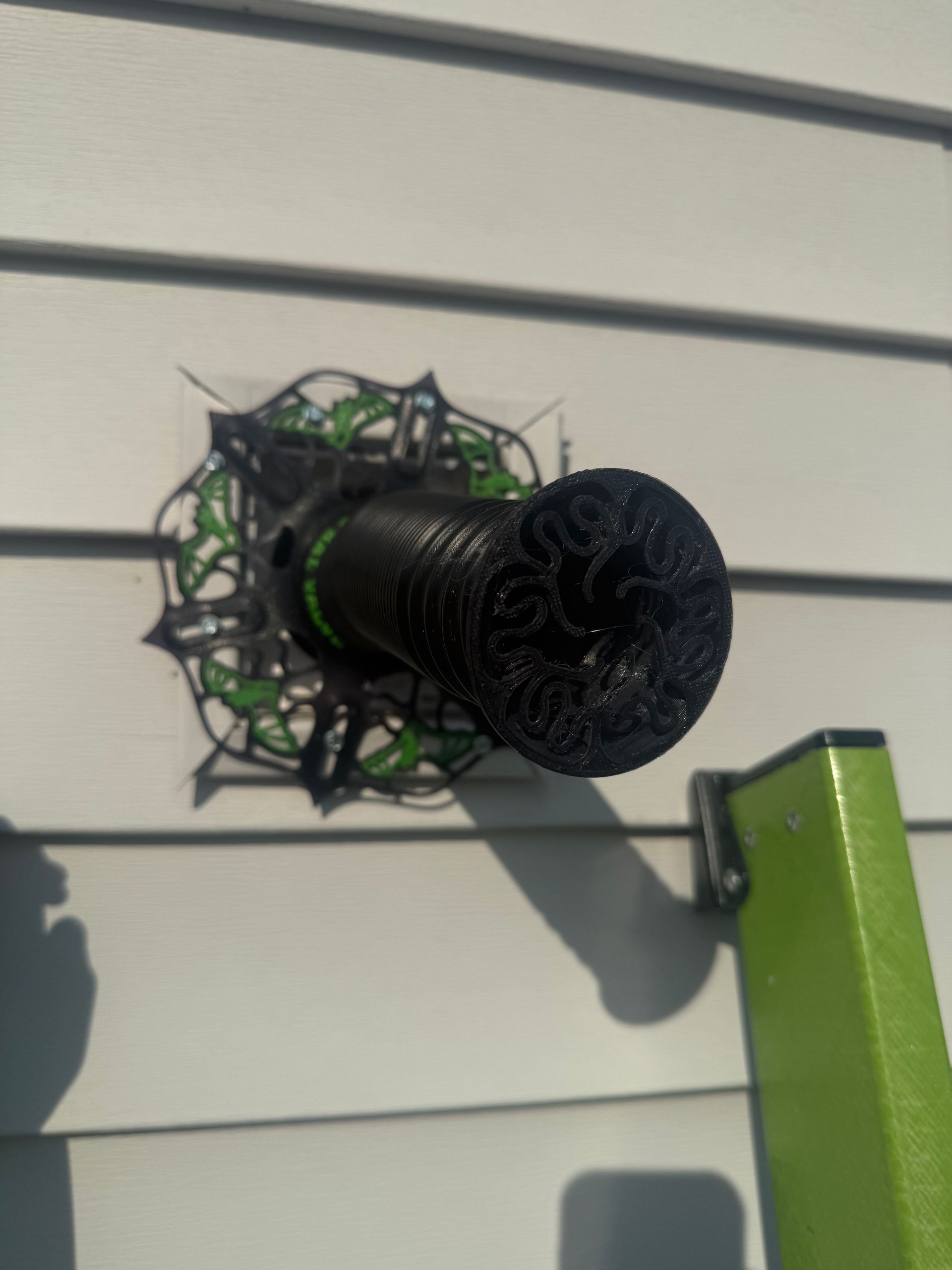 Black decorative vent mounted on beige siding with a green edge in the corner
