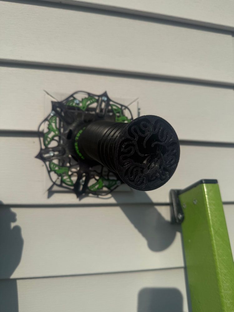 Black outdoor wall light mounted on beige siding beside a green post
