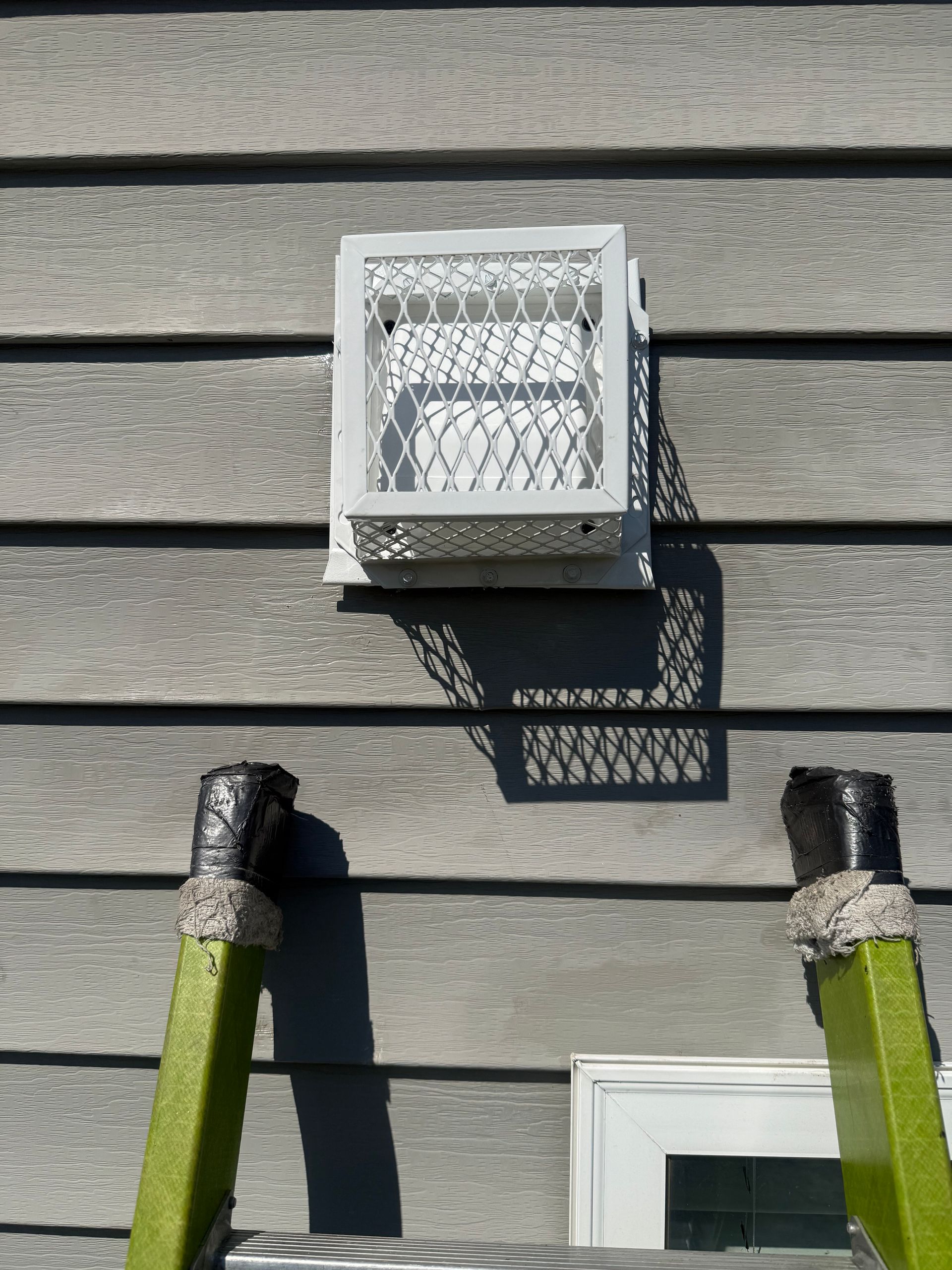 White vent on gray siding, with two green hoses below and a shadow cast to the right.