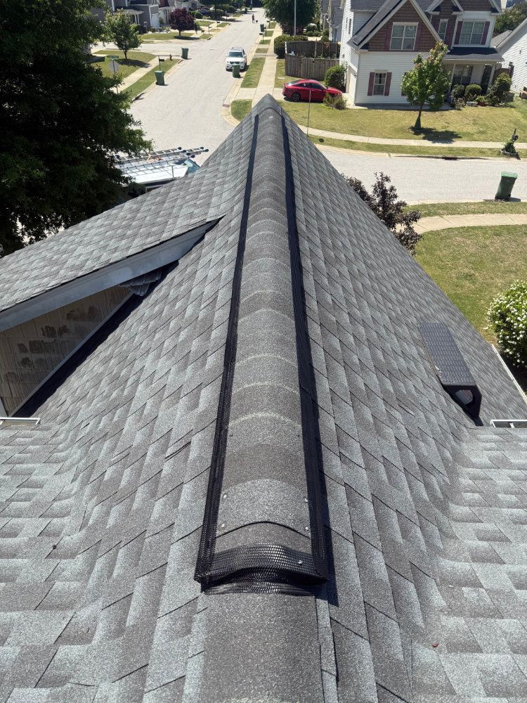 Aerial view of a gray shingled house roof with a central ridge vent in a suburban neighborhood.