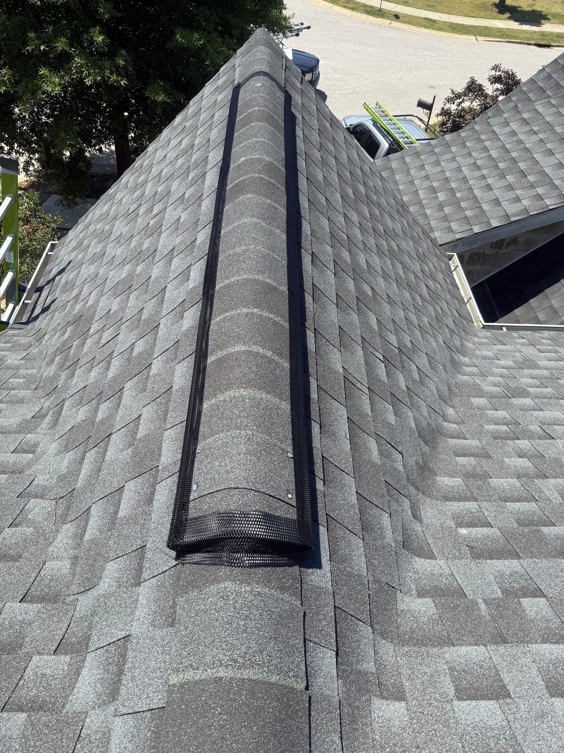 Roof ridge vent on a gray shingle roof, viewed from above with trees and driveway in the background