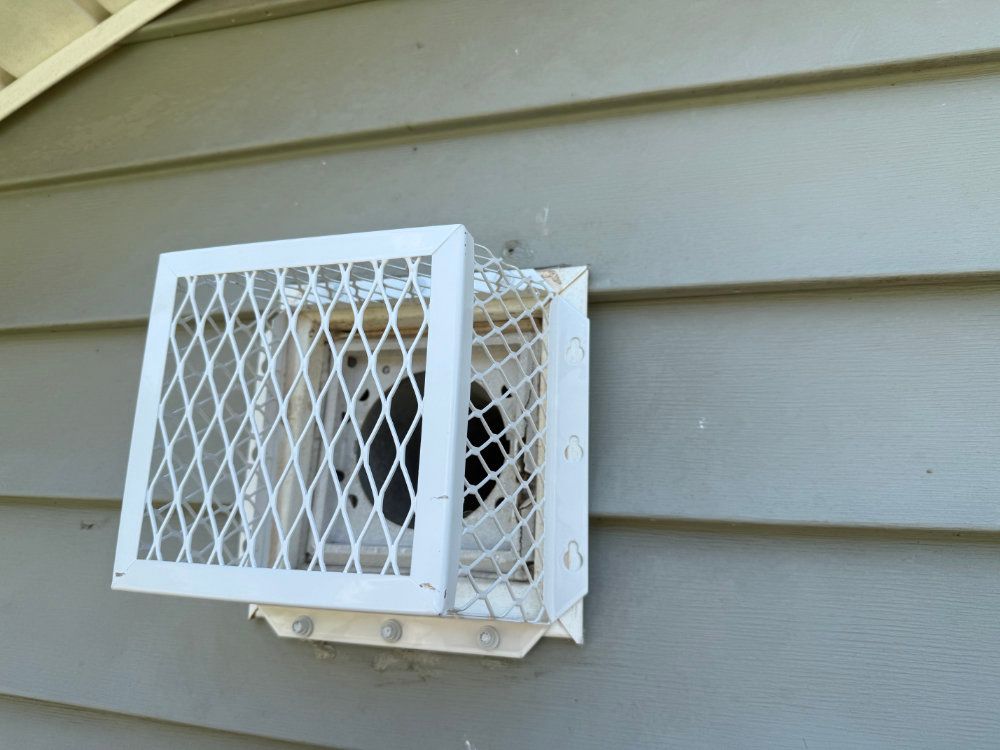 White metal vent cover mounted on beige siding, partially open with a diamond mesh grille.