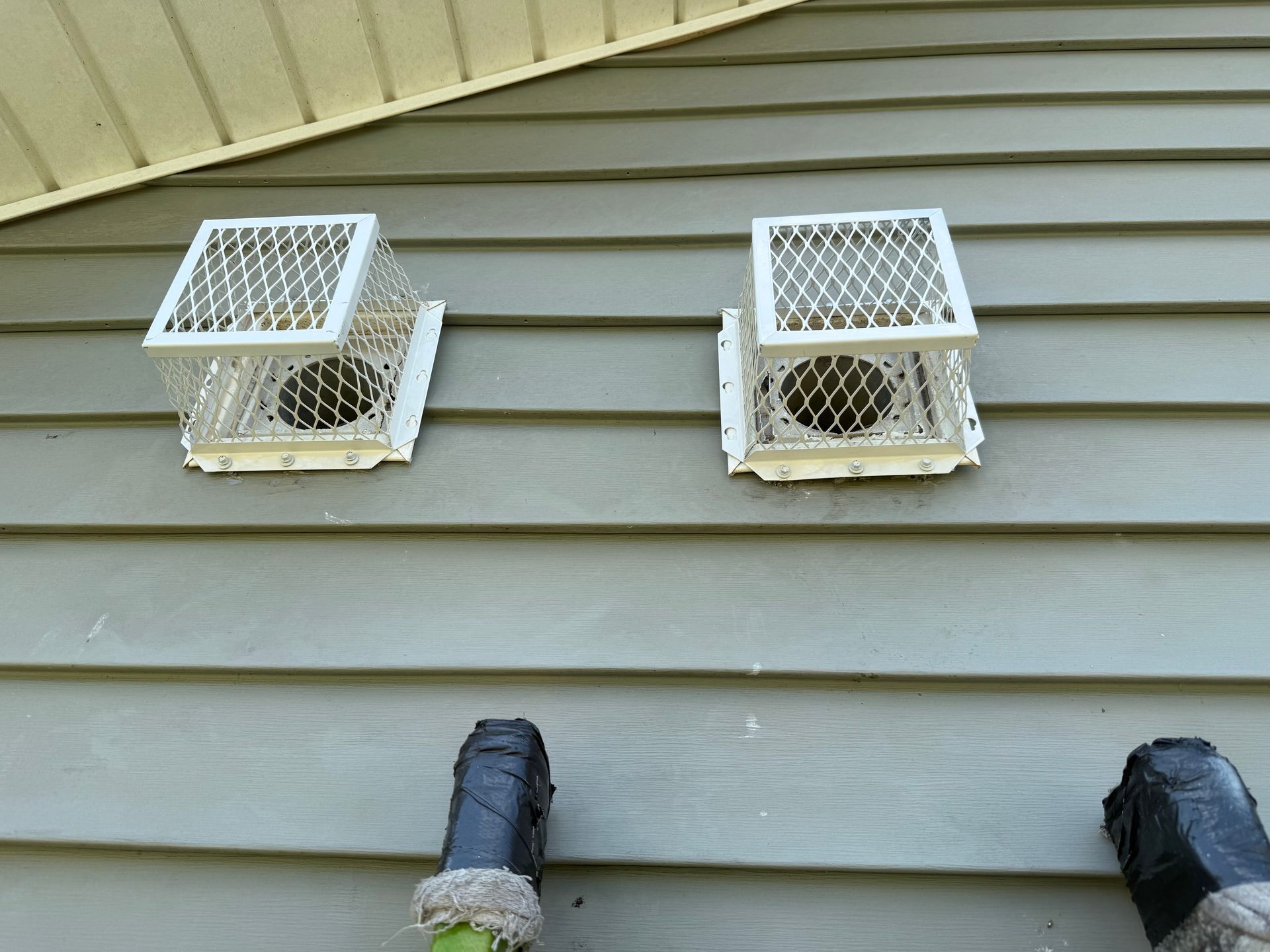 Two white wall vents with open covers on gray house siding, with two capped pipes below