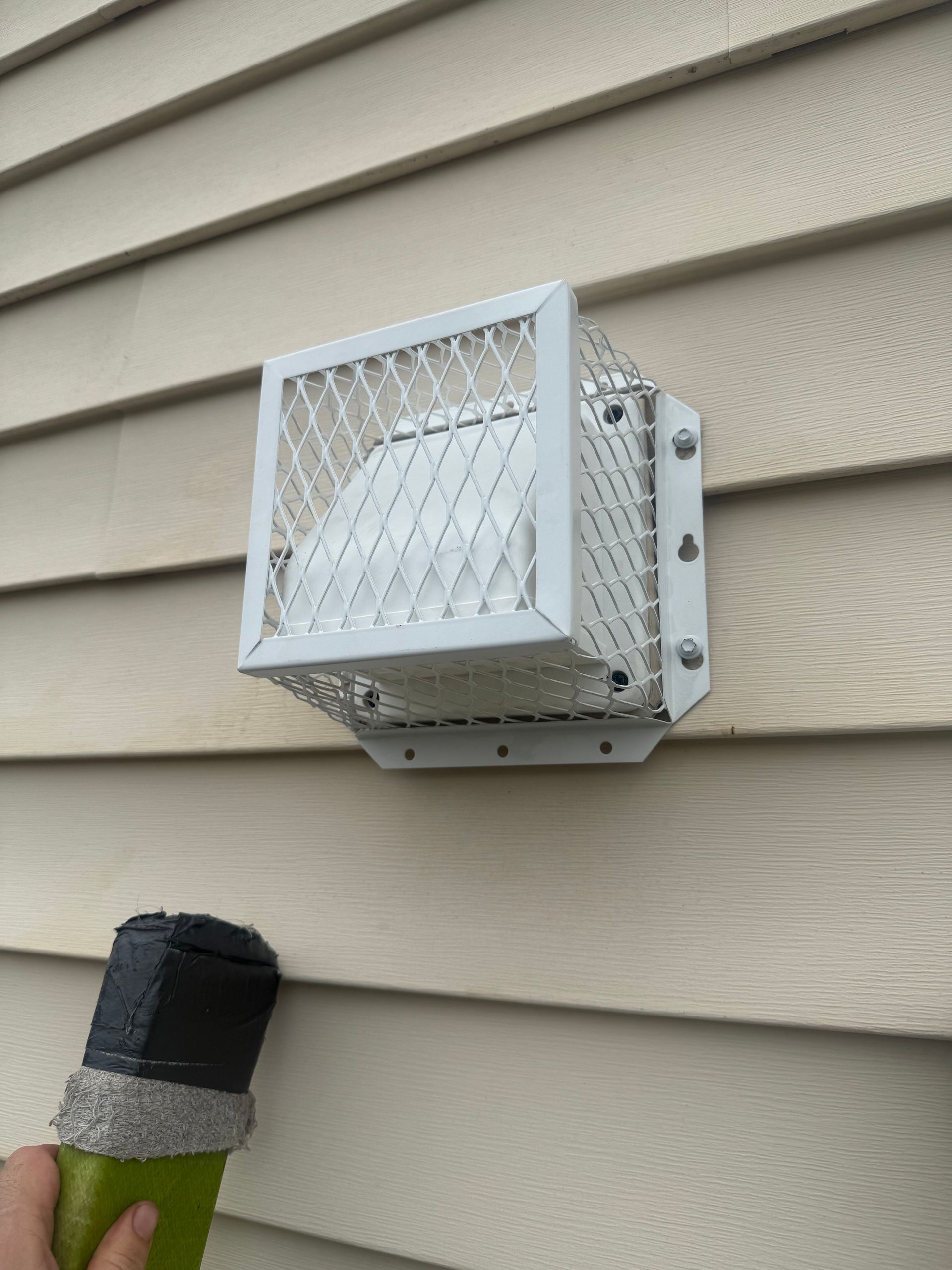 White outdoor security light on beige house siding above a black vent pipe