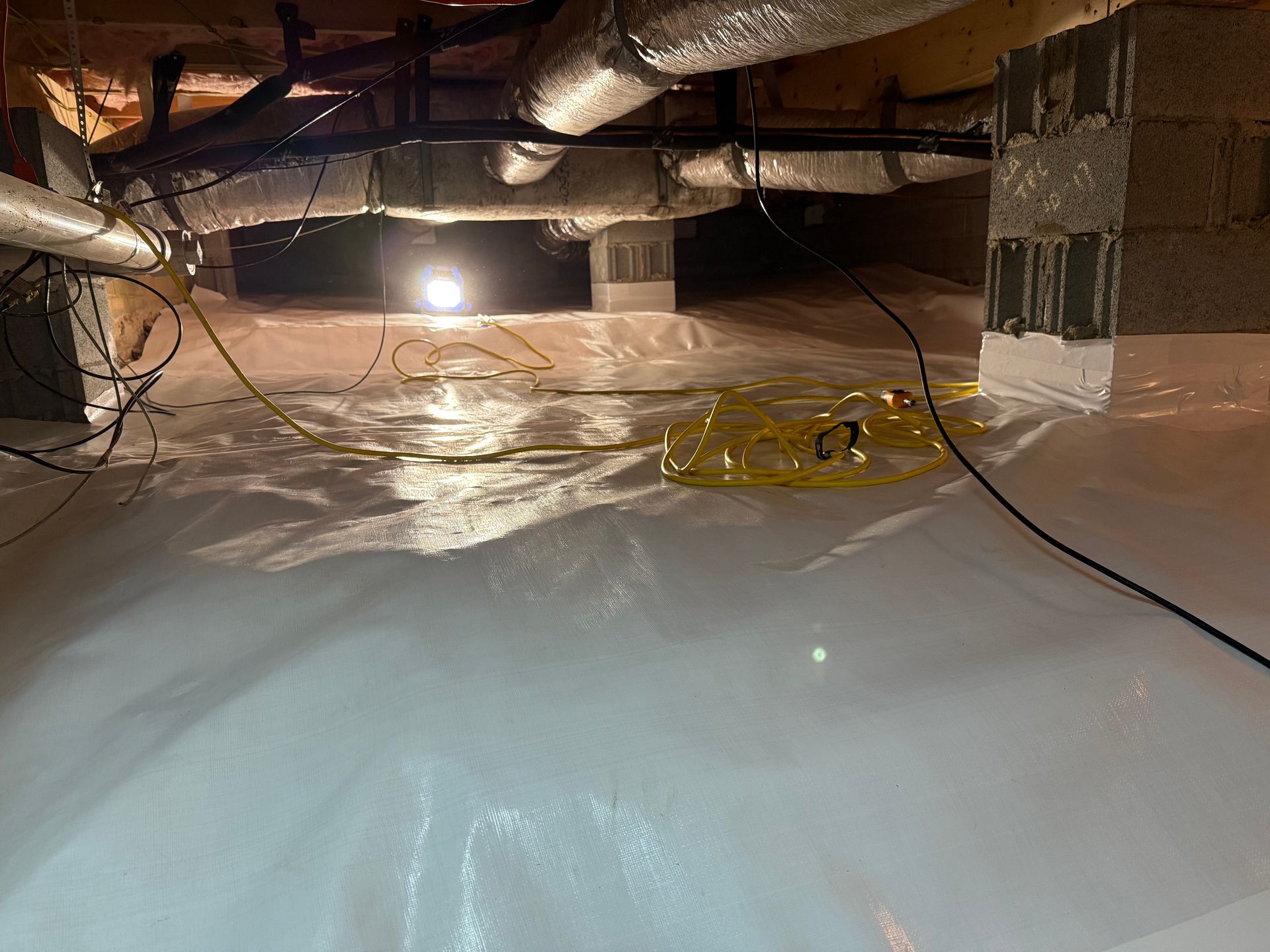 Crawlspace with plastic vapor barrier, exposed ducts, wiring, and a bright work light under floor joists