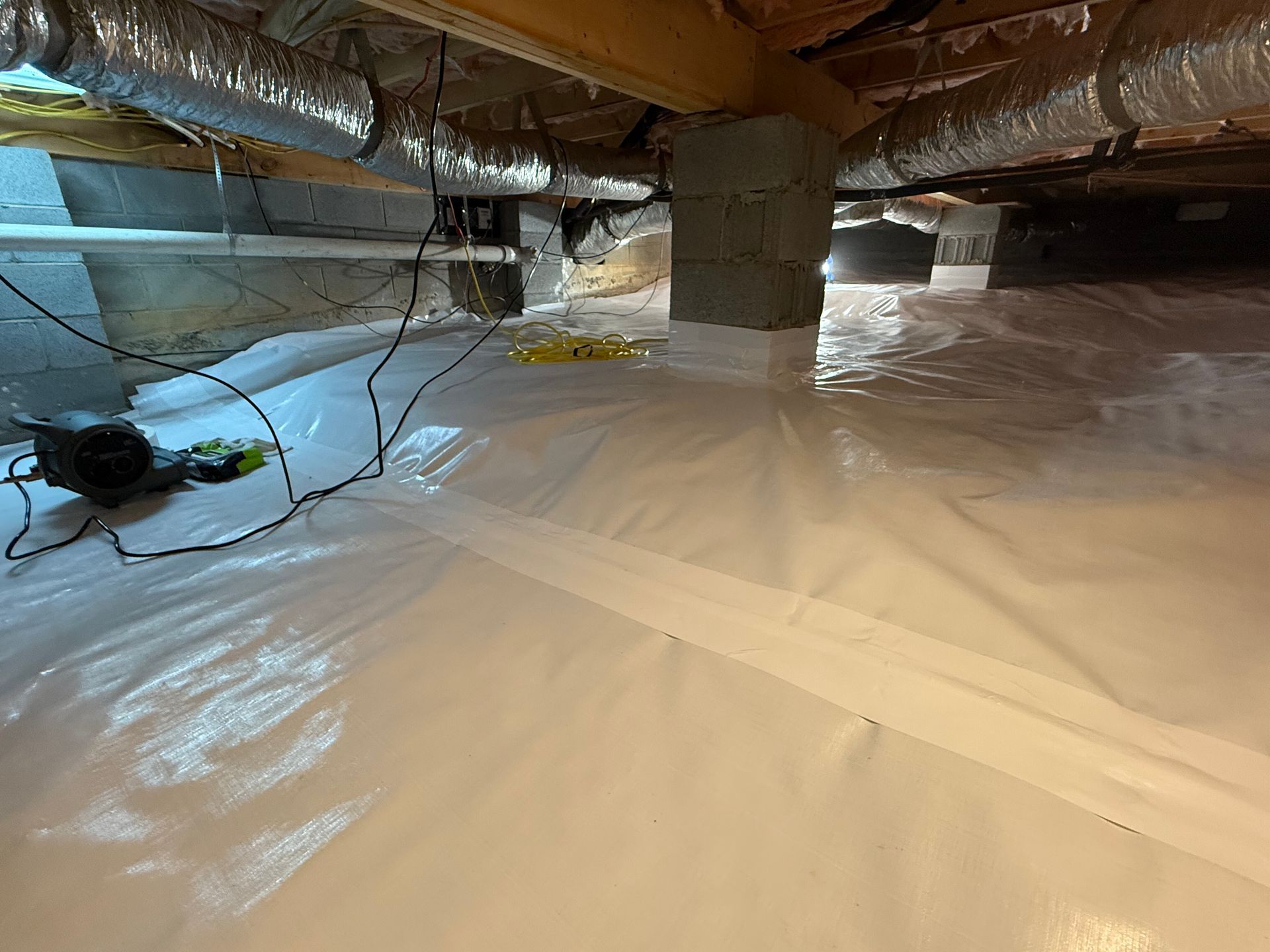 Crawlspace under house with white plastic vapor barrier, exposed insulation, and a black device on the floor
